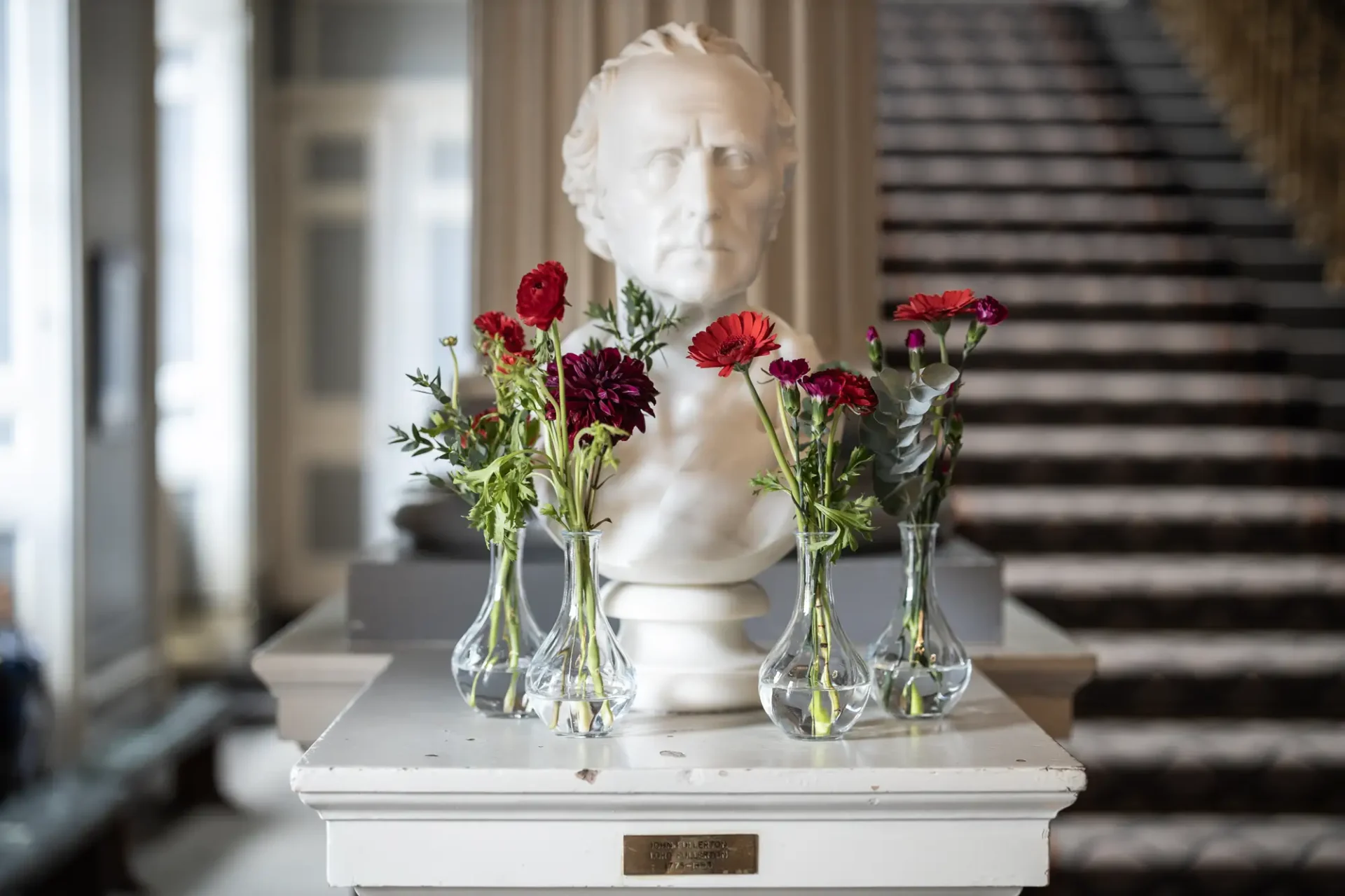 A marble bust of a man displayed on a pedestal, flanked by small vases containing red and burgundy flowers in an elegant room.