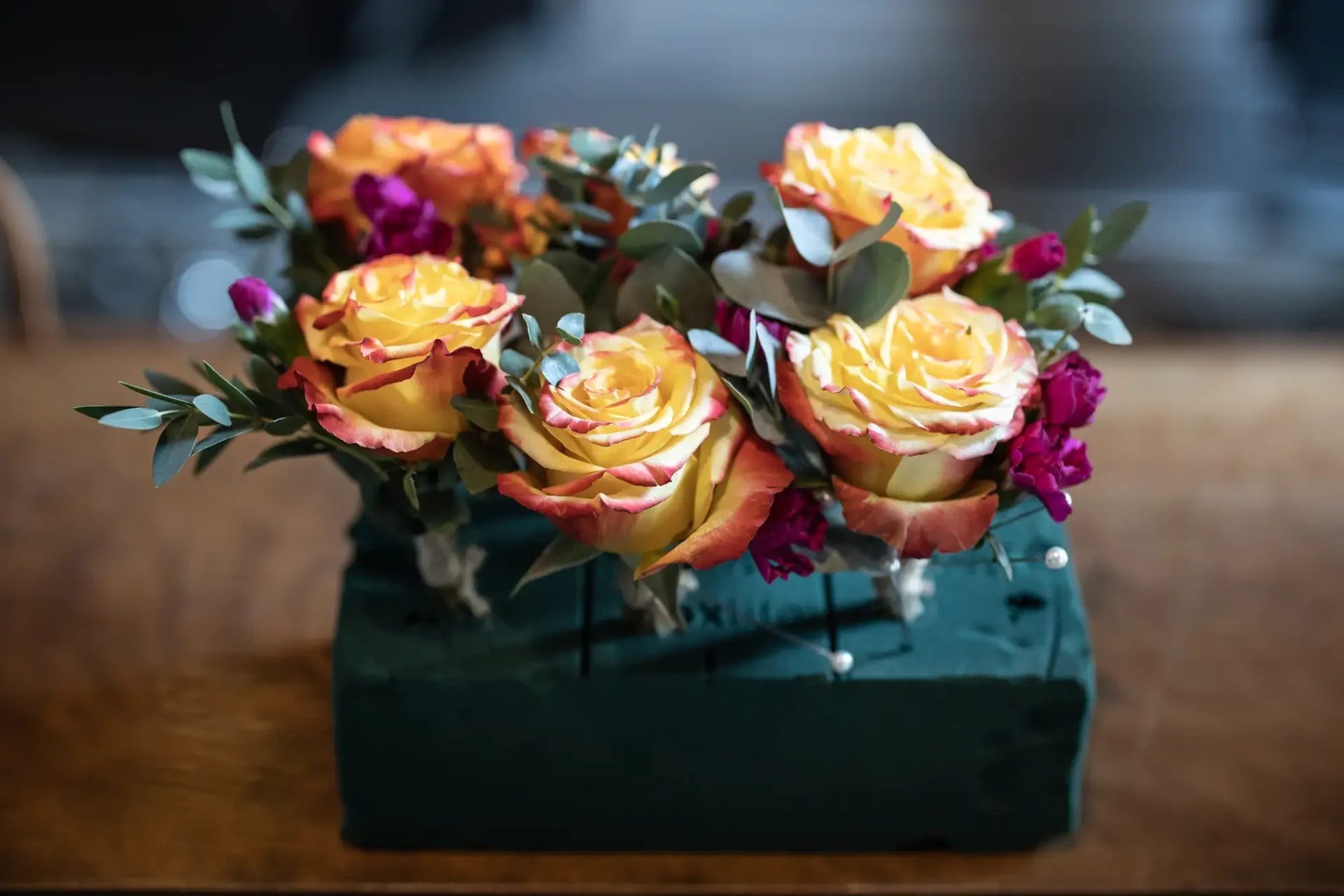 Floral arrangement featuring orange roses and purple flowers in a green square vase on a wooden table.
