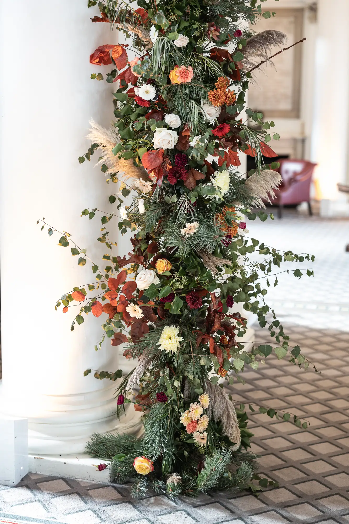 A slim, vertical christmas tree decorated with a mix of red, white, and yellow flowers, along with green leaves and feathers, set against a white pillar.