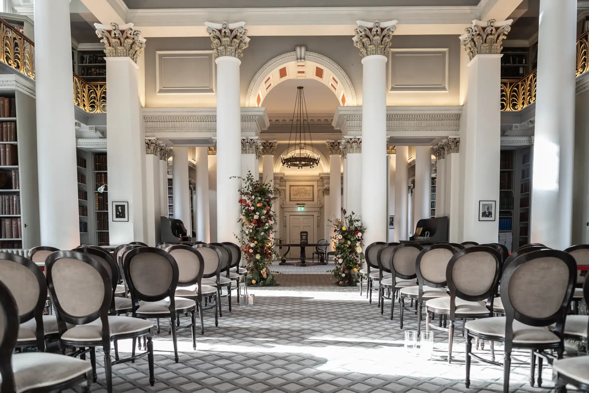 Elegant hall arranged for a wedding with rows of chairs and floral decorations, featuring classical architecture and large windows.