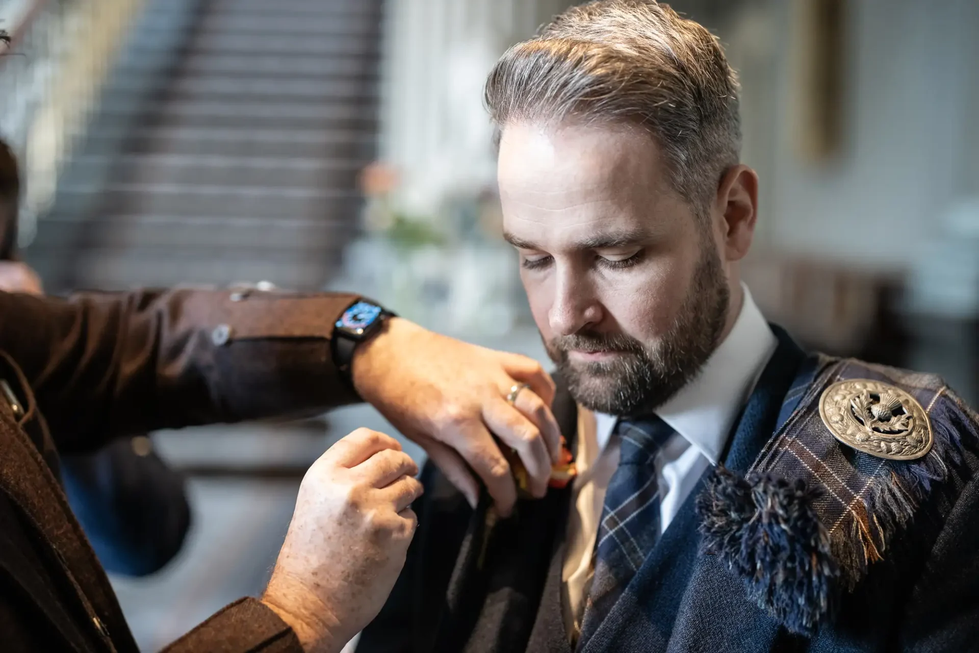 A man in a formal kilt outfit getting assistance with his cufflinks indoors.