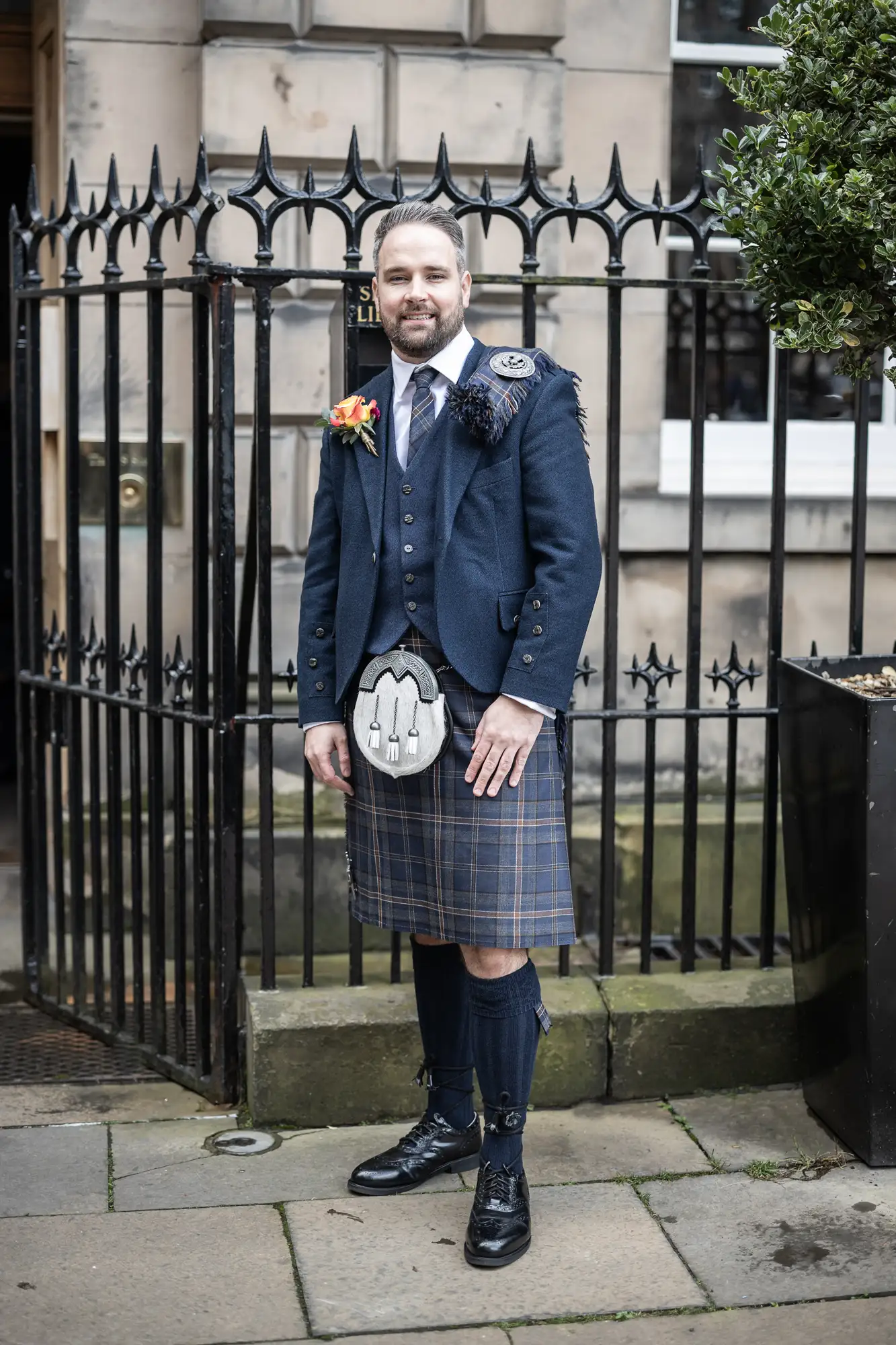 Man in traditional scottish attire, including a kilt and sporran, stands smiling by a black iron fence with a boutonniere on his jacket.