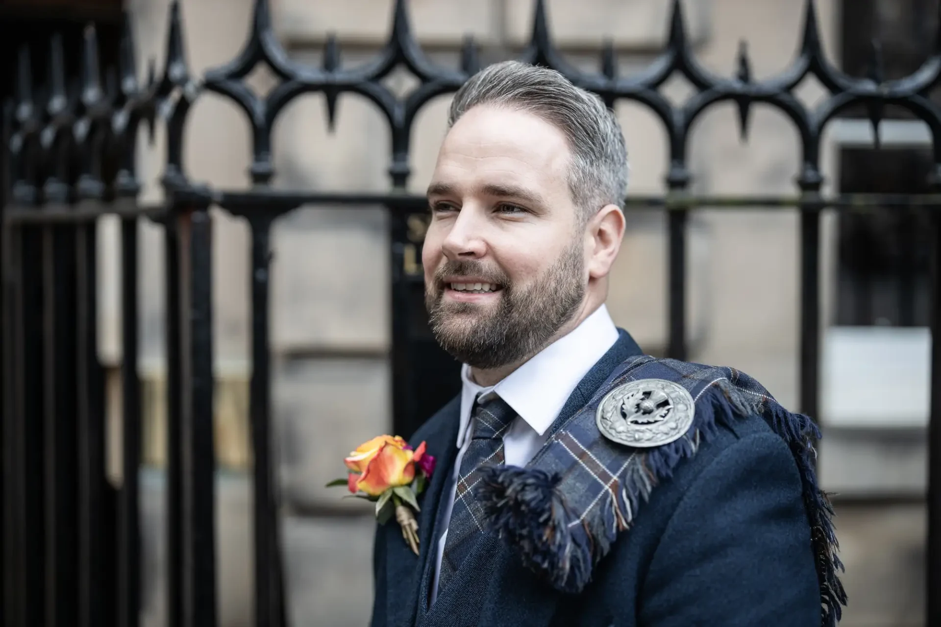 Man in a traditional kilt and tweed jacket, smiling outside a building with iron fence, wearing a boutonniere.