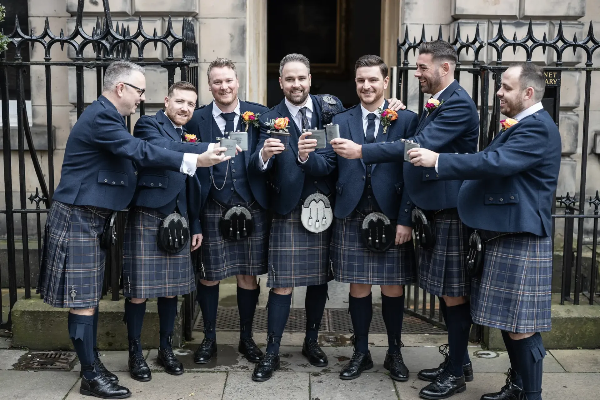 Six men in matching tartan kilts and jackets, toasting with drinks, celebrating outdoors near a building with iron fences.