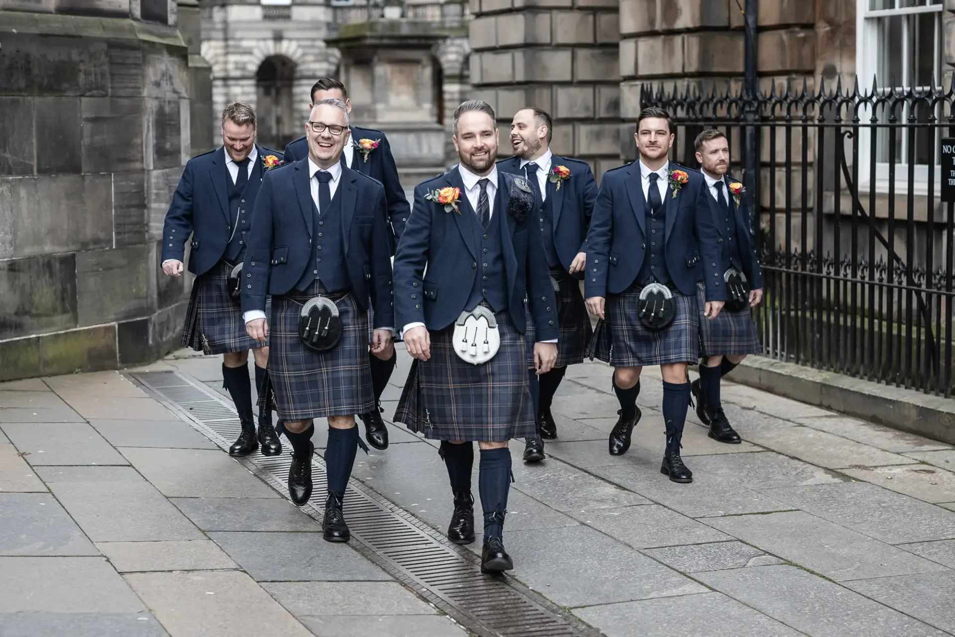 A group of six smiling men in traditional scottish attire, including kilts and sporran, walking together on a city street.