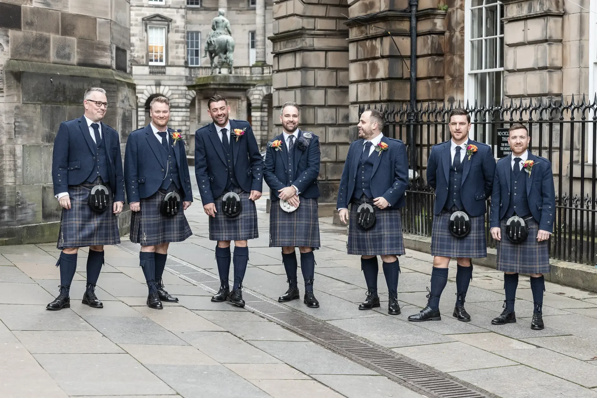 Seven men in matching tartan kilts and jackets smiling and holding their hats, standing on a cobblestone street.