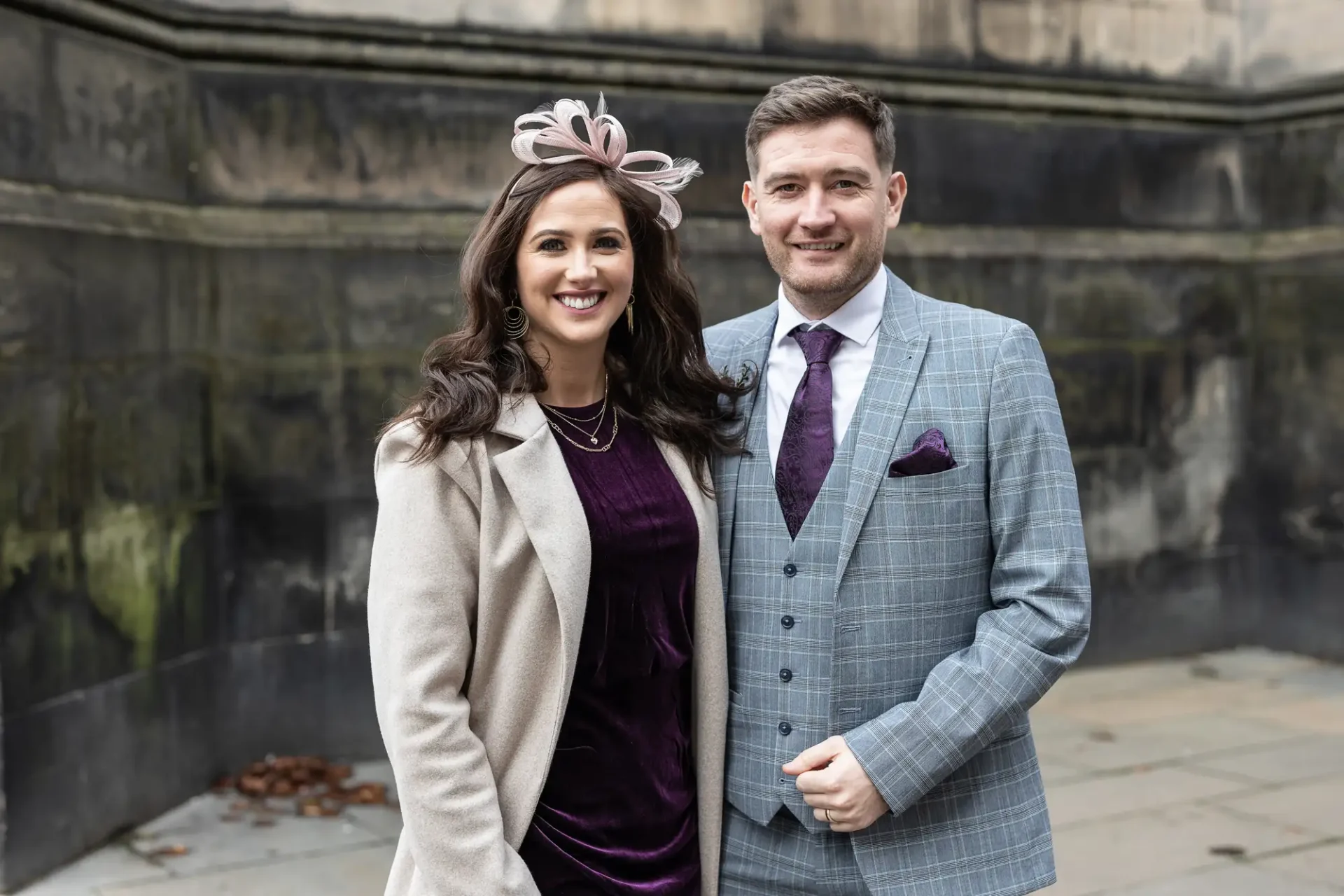 A man and woman, dressed formally, smiling outdoors; the man wears a plaid suit and the woman a dress with a coat and fascinator.