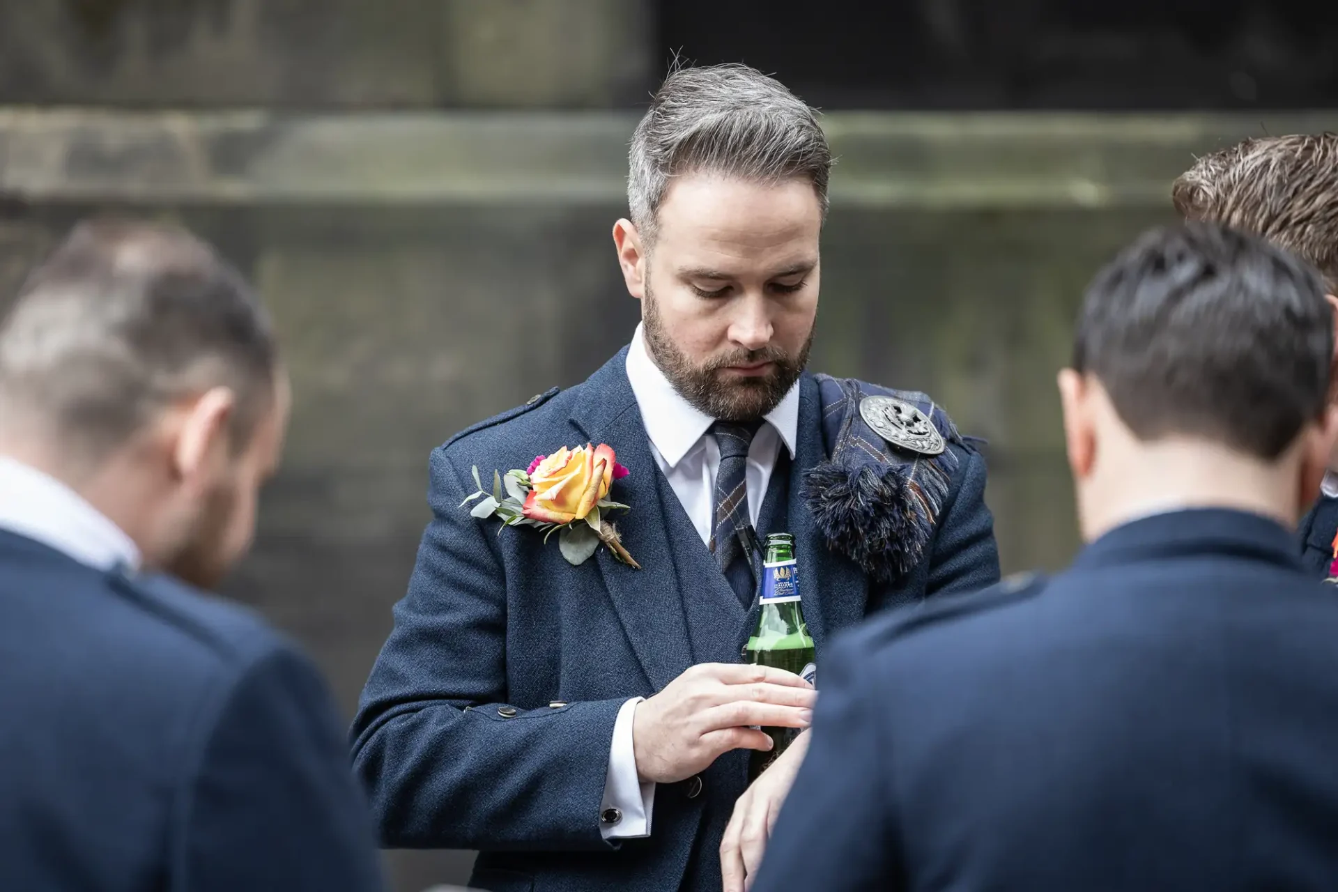 A man in a formal suit with a boutonniere checks his watch, standing among other men in a conversational outdoor setting.