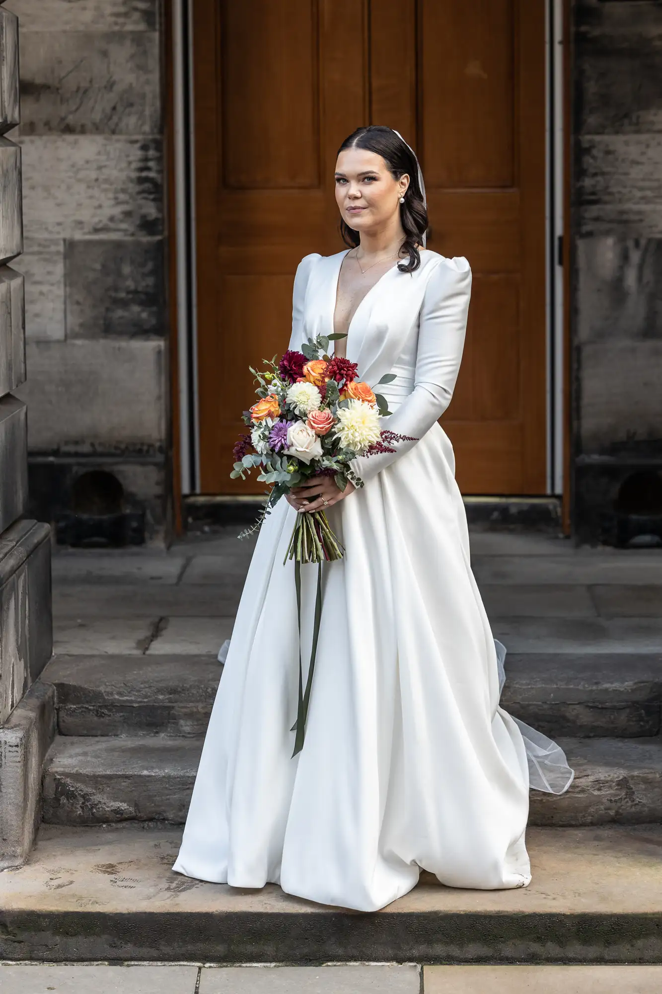 A bride in a white long-sleeve gown holds a colorful bouquet, standing in front of a dark wooden door.