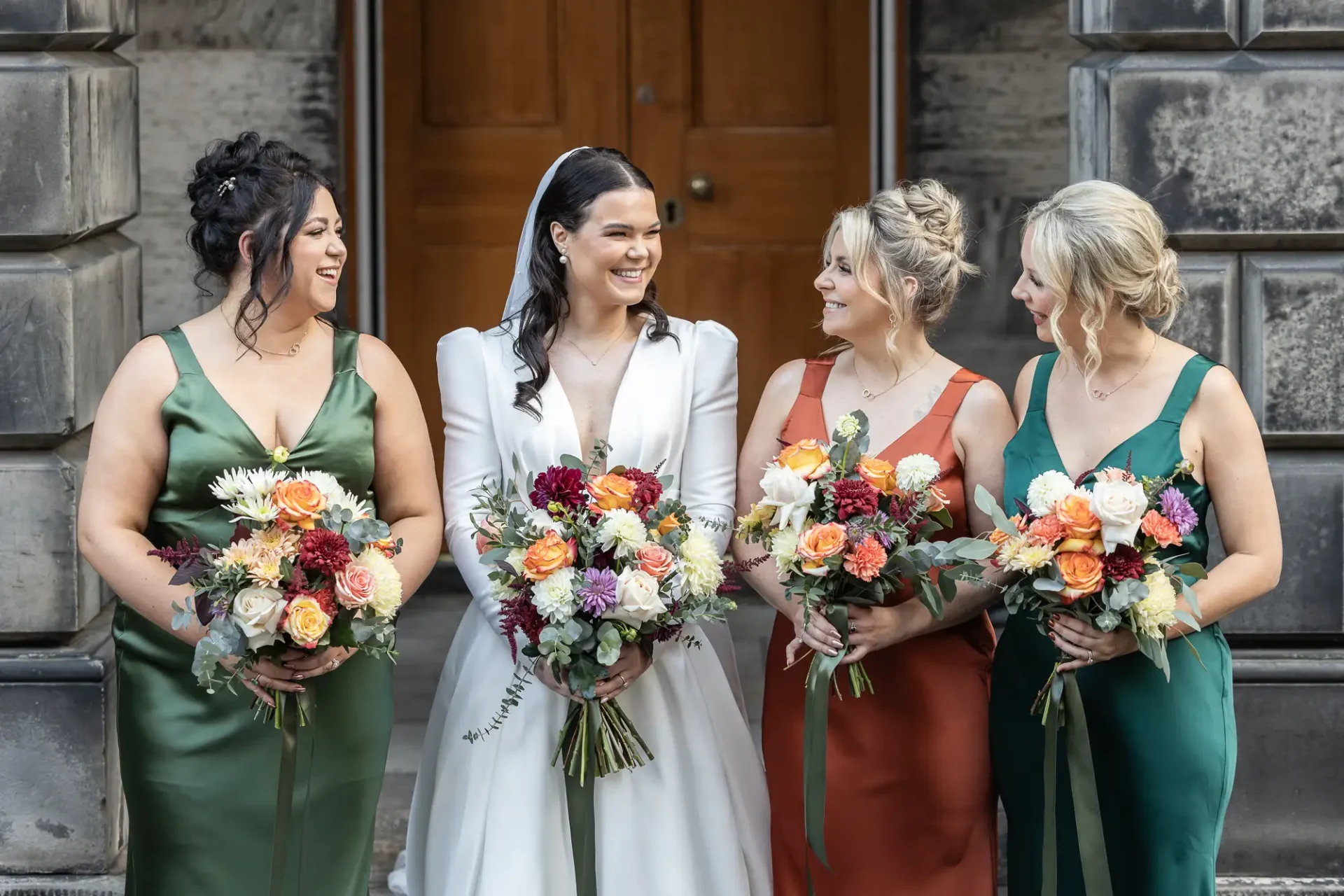 Bride in white dress smiling with three bridesmaids in green and teal dresses, holding colorful bouquets, standing outside a stone building.