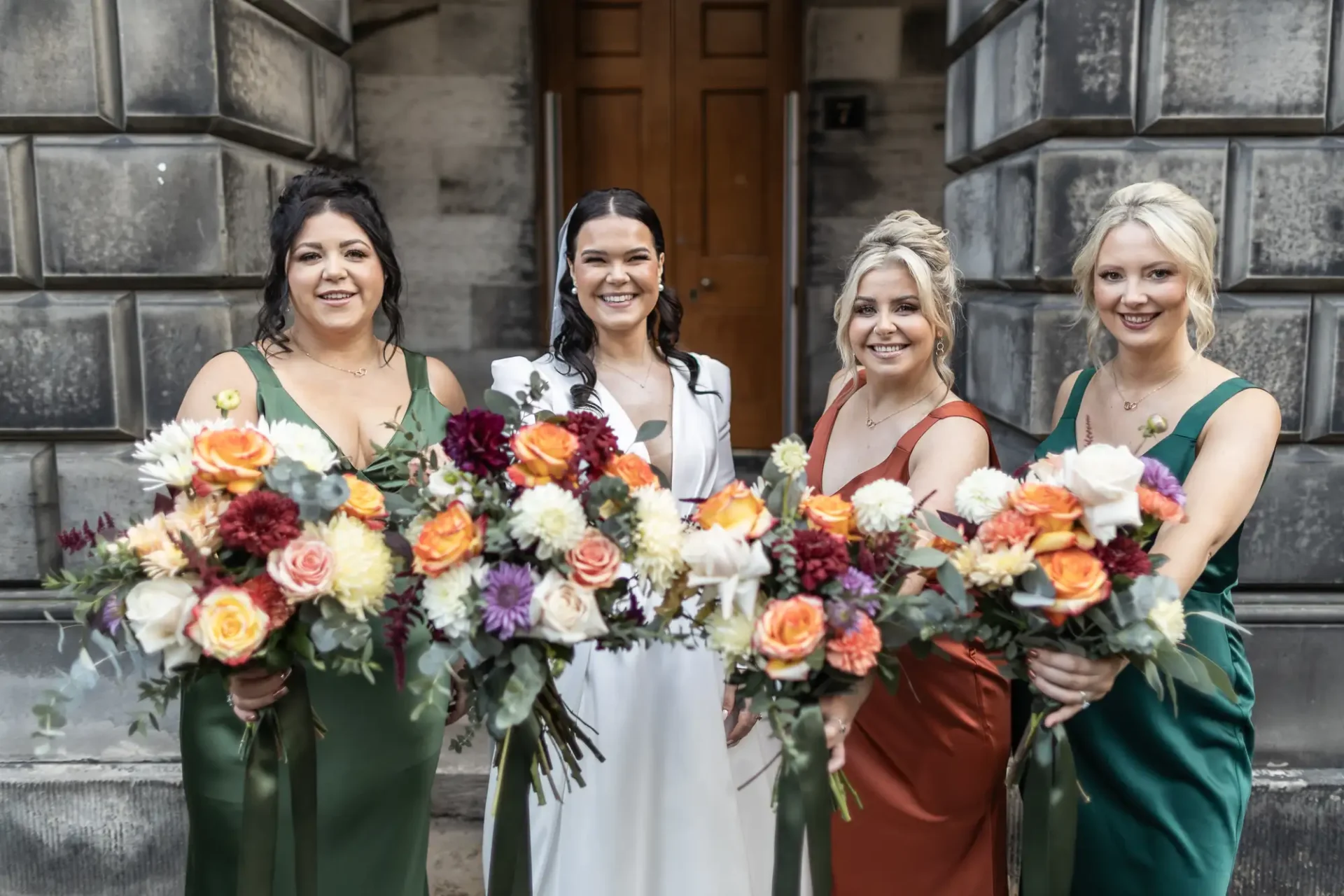 A bride and three bridesmaids posing with large, colorful bouquets in front of a stone building.
