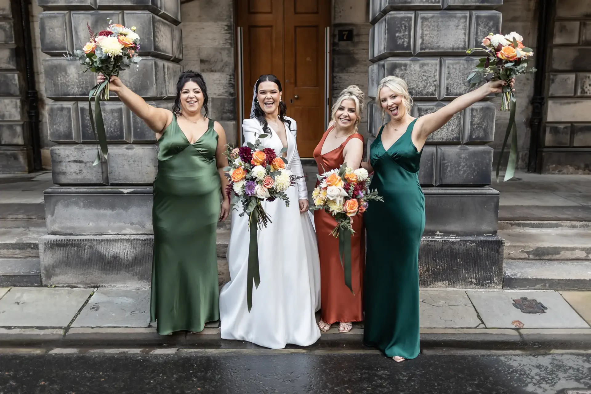Four women in elegant dresses, holding bouquets, joyfully posing outside a stone building. two wear green and two in white and rust.