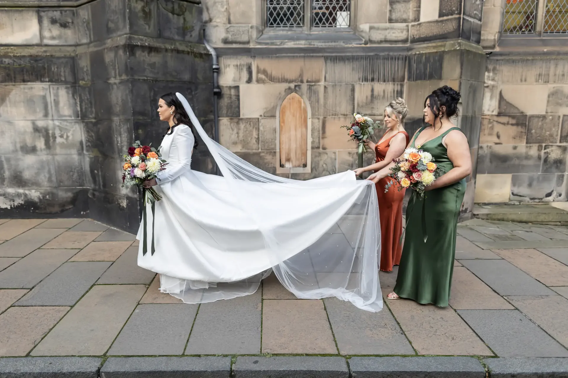 A bride in a white dress with a flowing veil, flanked by three bridesmaids in green and peach dresses, holding bouquets, outside a stone building.