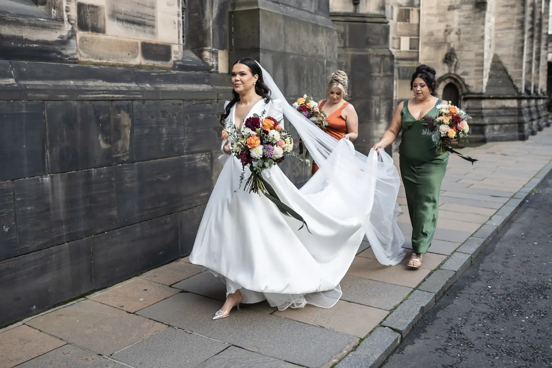 A bride in a white dress leads two bridesmaids carrying bouquets down a city street; one bridesmaid is adjusting the bride's train.