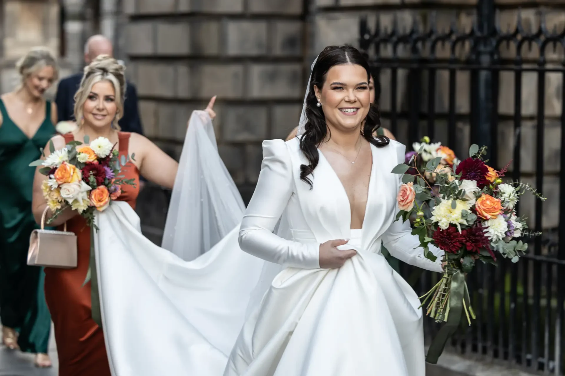 A radiant bride with a bouquet smiling as she walks, accompanied by two bridesmaids holding her dress train.