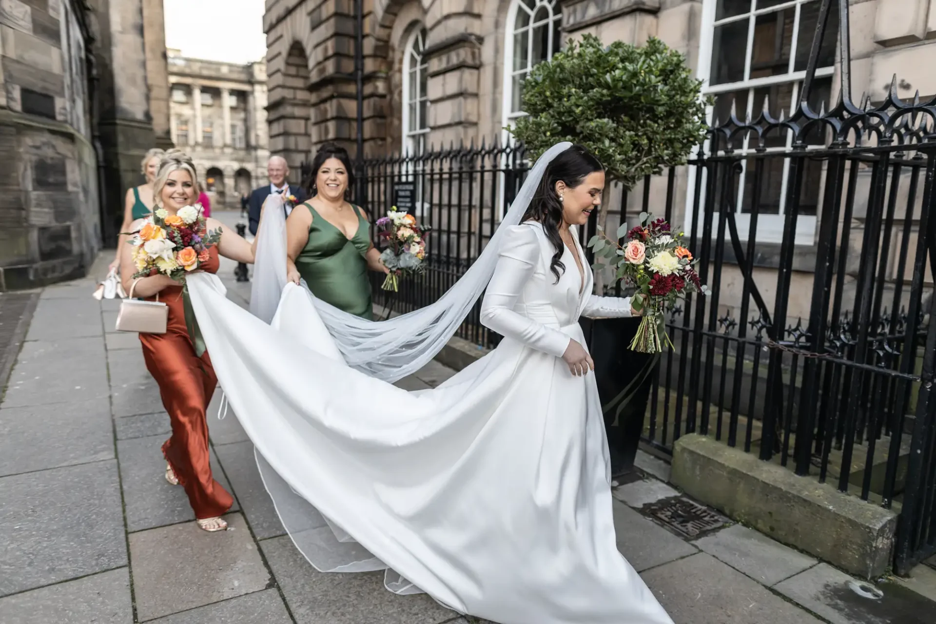 A bride in a flowing white gown walks with three bridesmaids holding colorful bouquets on a city street.
