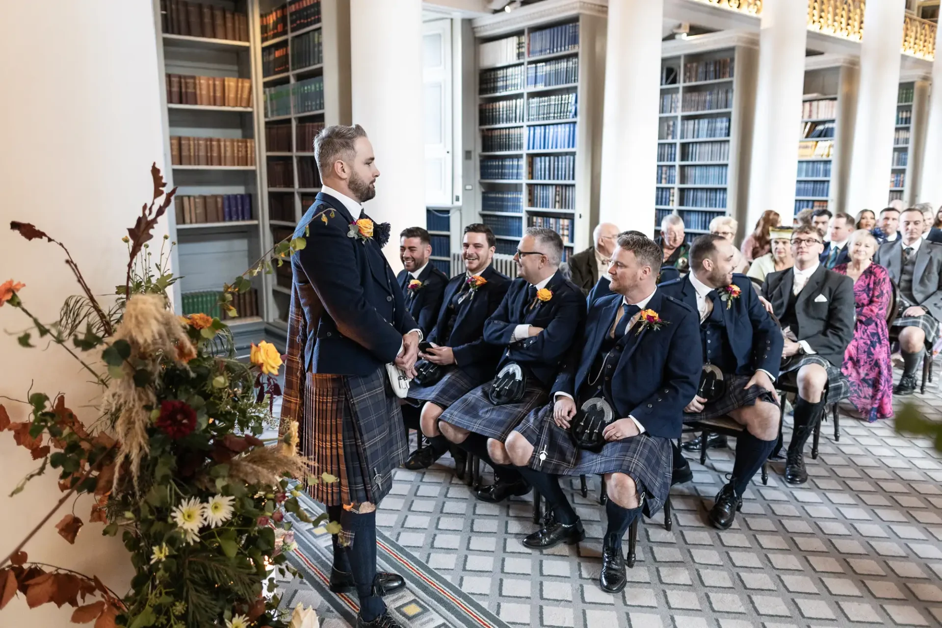A groom in a kilt smiles at seated wedding guests, also mostly in kilts, in an elegant library setting adorned with floral decorations.
