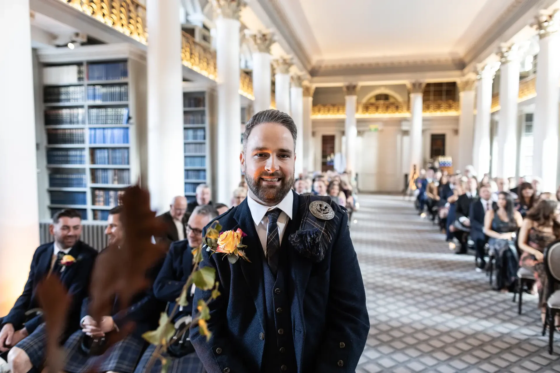 A smiling groom in a kilt walking down the aisle in a library setting with seated guests blurred in the background.