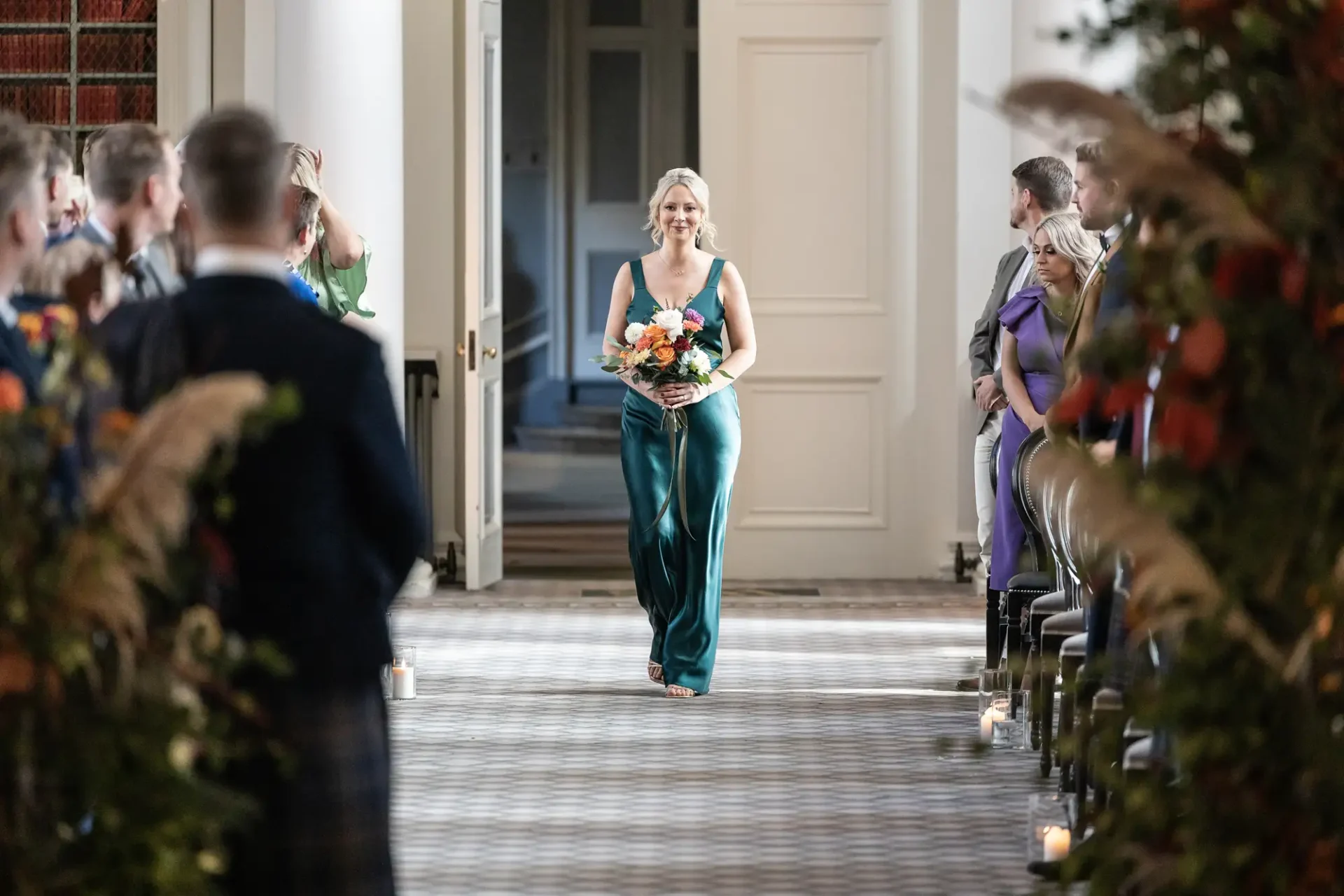 A woman in a teal dress walks down the aisle holding a bouquet, smiling at guests during a wedding ceremony.