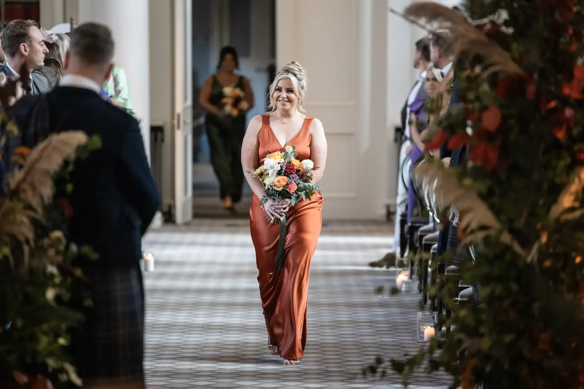 A woman in an orange dress smiles as she walks down an aisle holding a bouquet, flanked by seated guests and plant decorations.