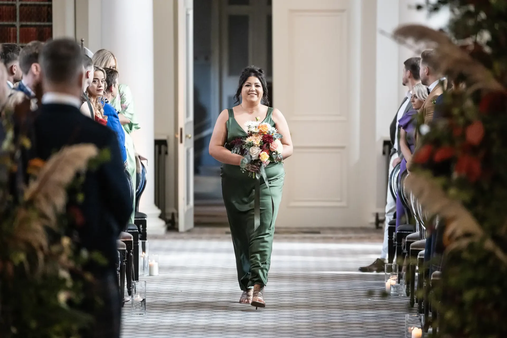 A woman in a green jumpsuit smiling as she walks down the aisle holding a bouquet, with guests seated on either side.