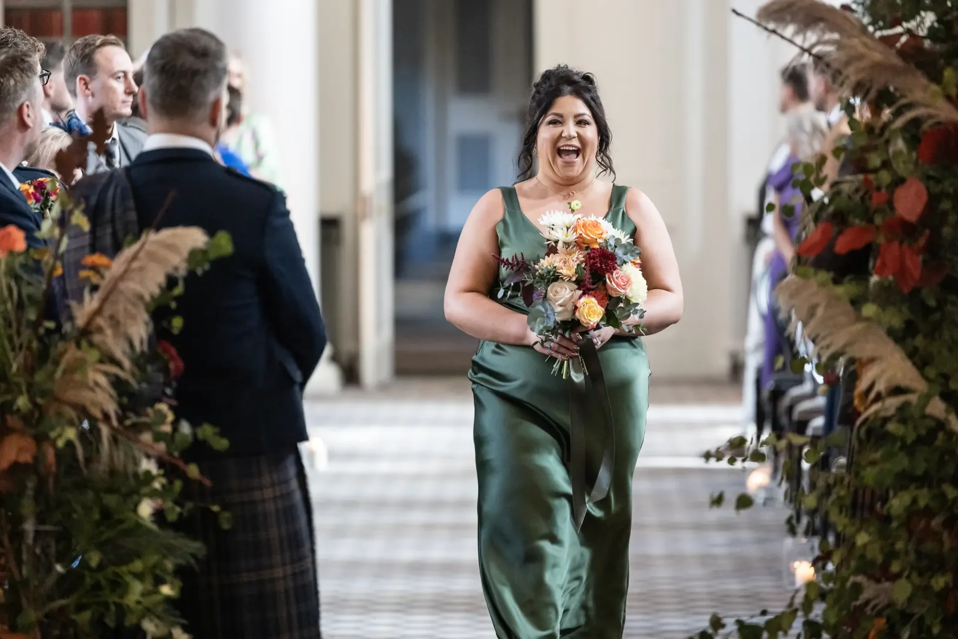 A joyful bridesmaid in a green dress walking down the aisle with a bouquet, laughing, as guests watch.