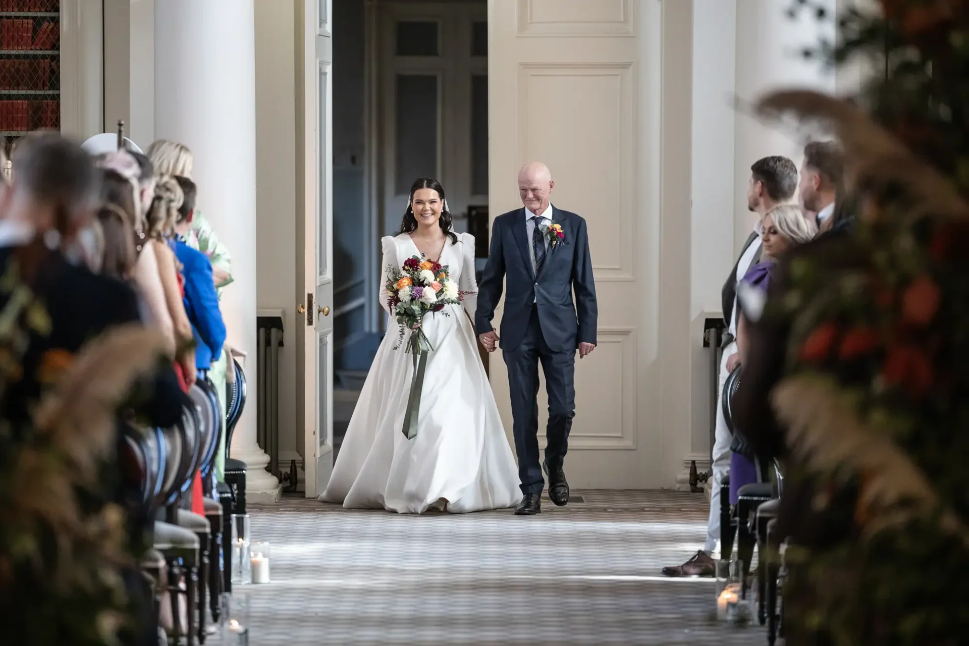 A bride and her father walk down the aisle, smiling, with seated guests on either side in a well-lit, elegant hallway.