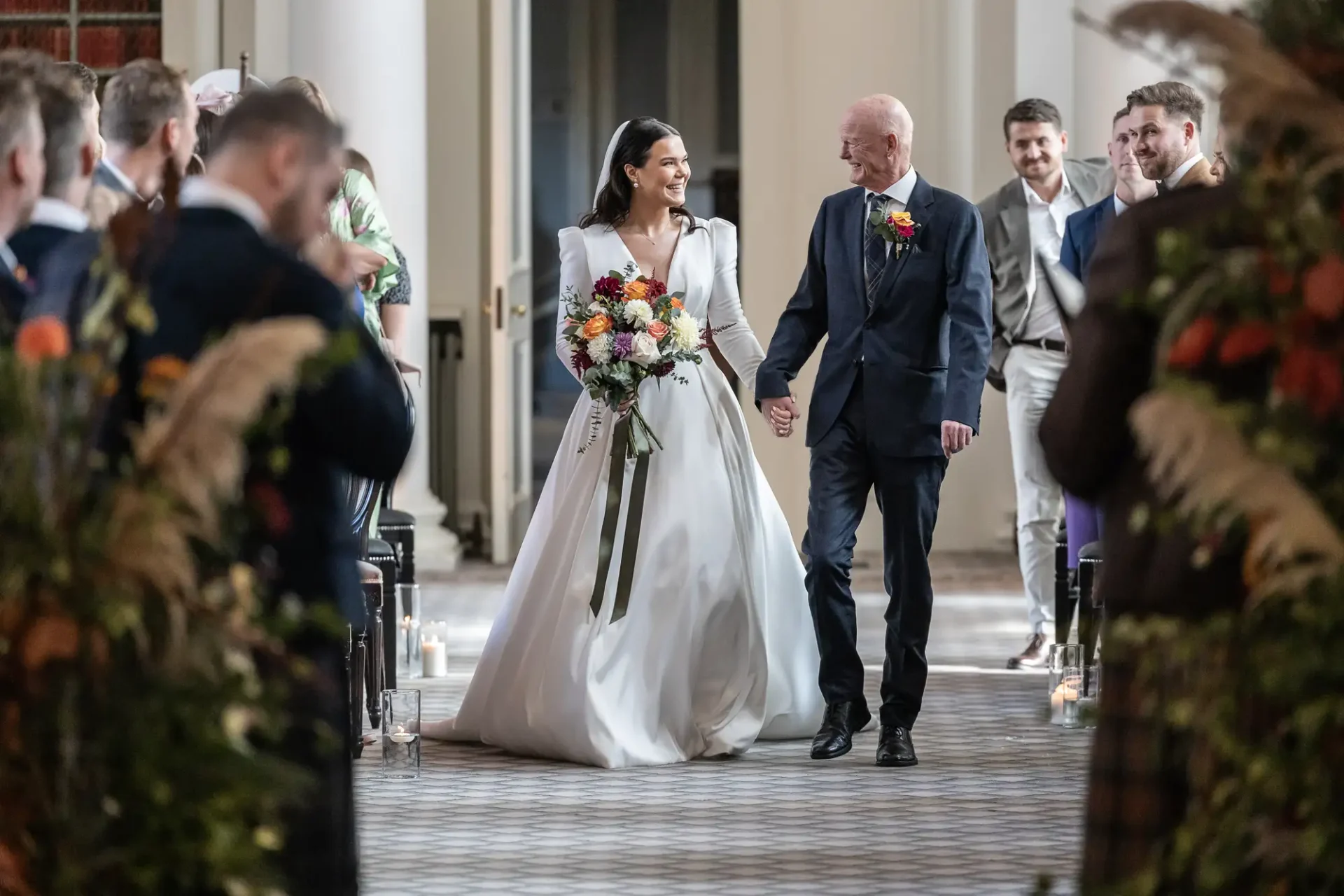A bride in a white dress walks down the aisle smiling, escorted by an older man in a suit, with guests standing on either side.