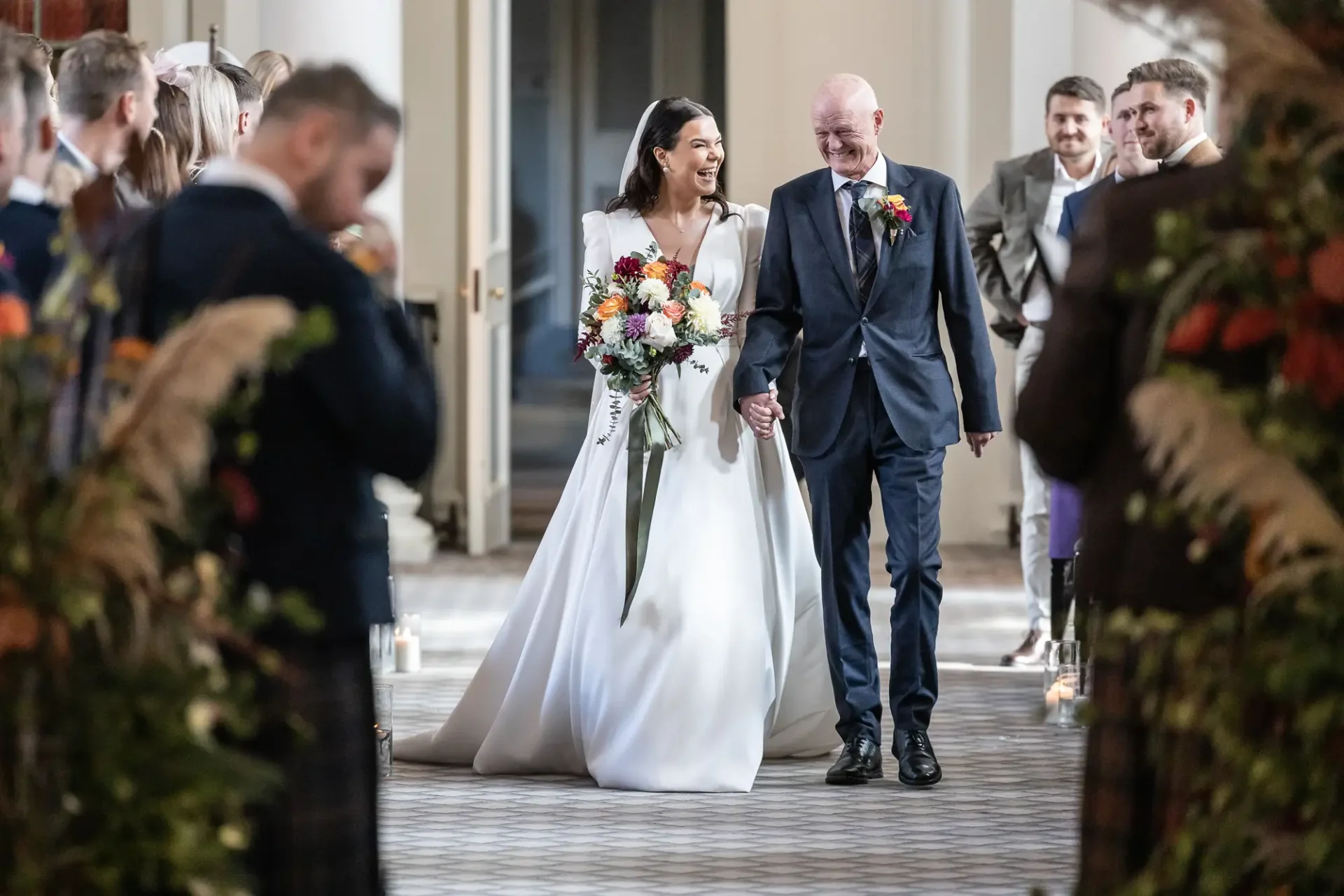 A bride and her father walk down the aisle, smiling, with guests on either side in a well-lit venue.