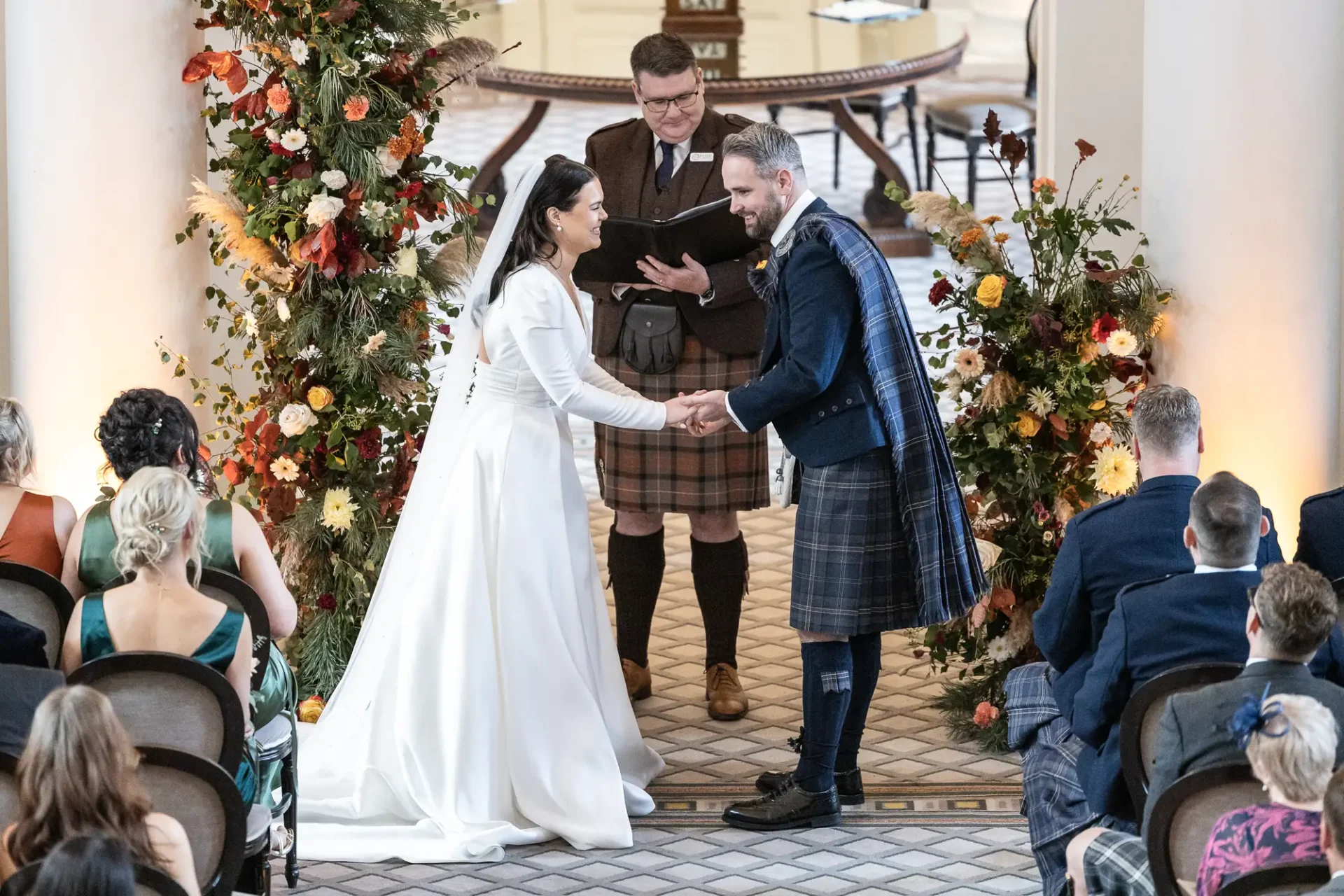A bride and groom holding hands at their wedding ceremony, surrounded by floral decorations and guests, with the groom wearing a kilt.
