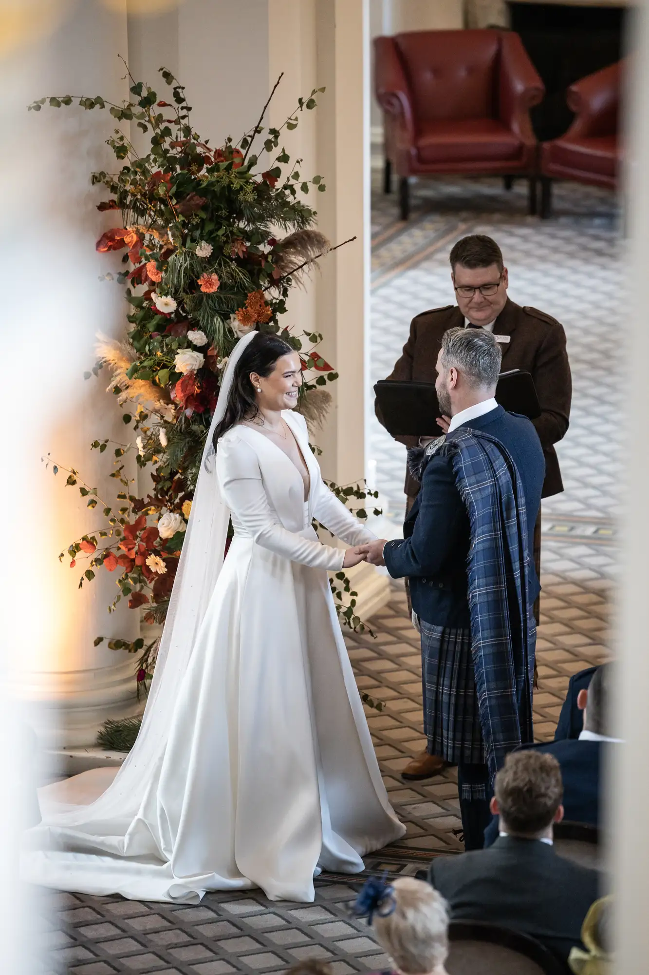 A bride in a white dress and a groom in a plaid kilt exchanging vows at their wedding ceremony, with an officiant and floral decorations in the background.
