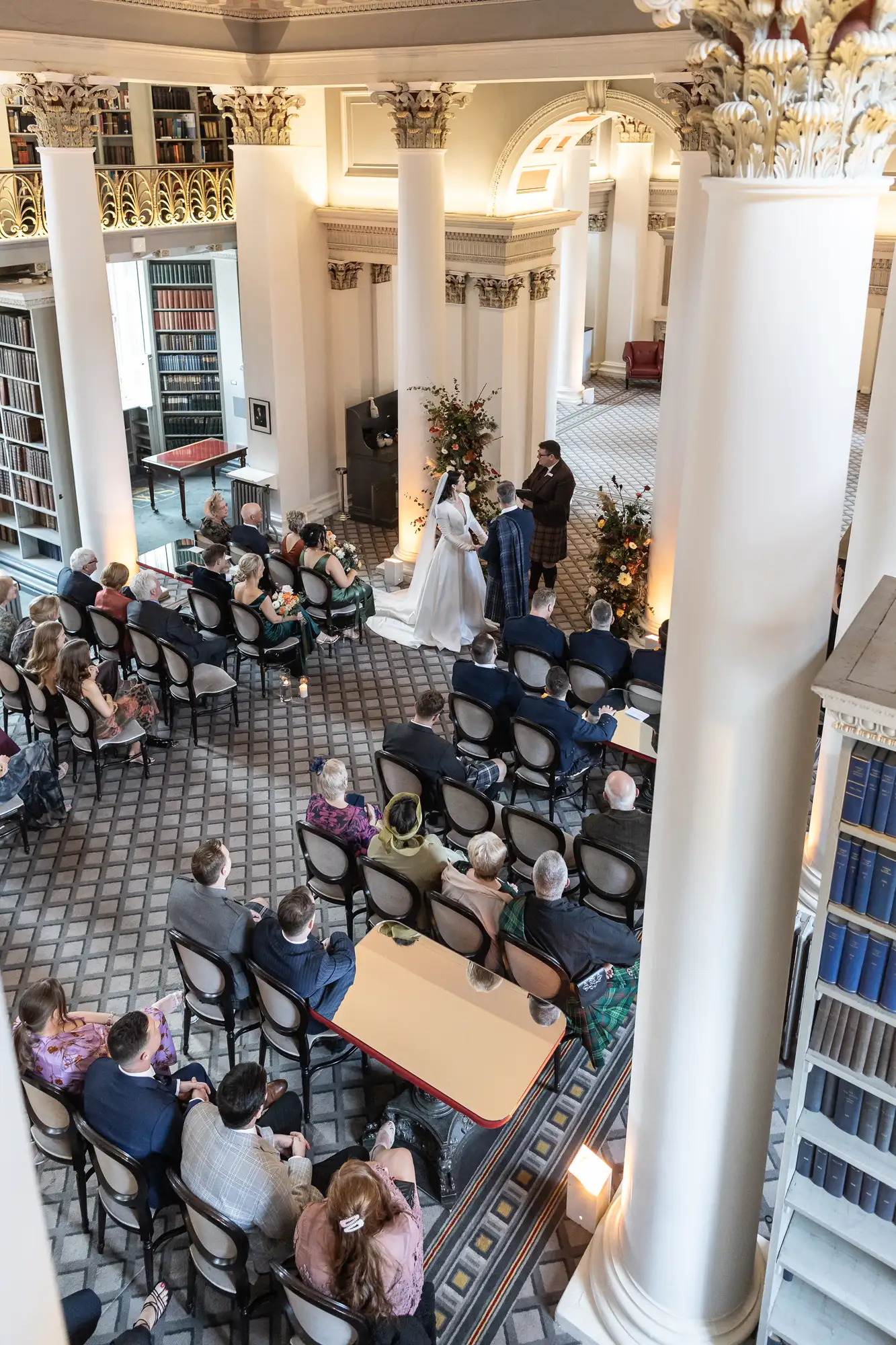 A wedding ceremony in an elegant hall with large columns, viewed from above, as guests watch the couple exchanging vows.