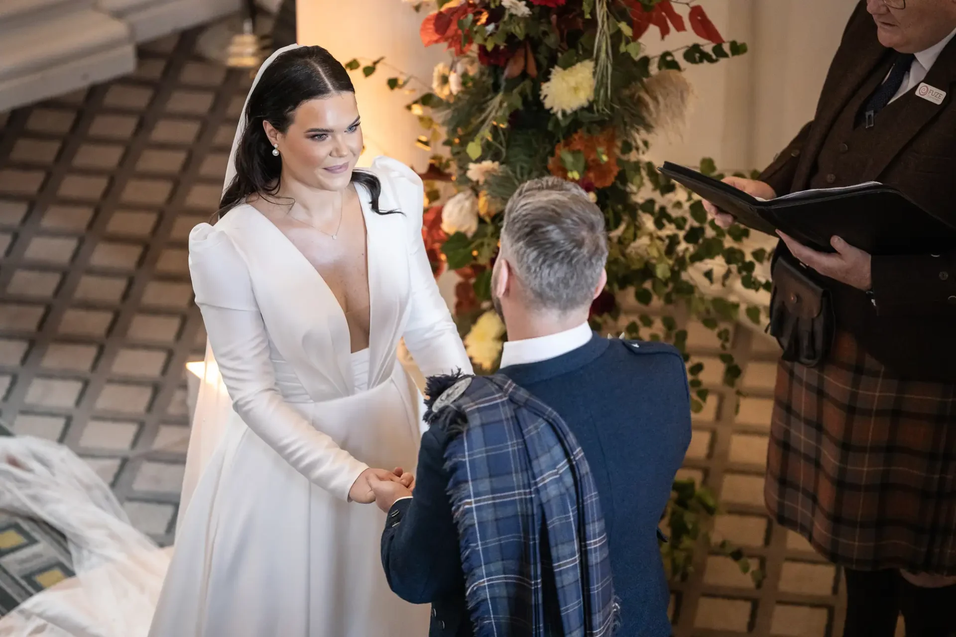 A bride in a white dress stands facing the groom in a tartan kilt during their wedding ceremony, with an officiant on the right.