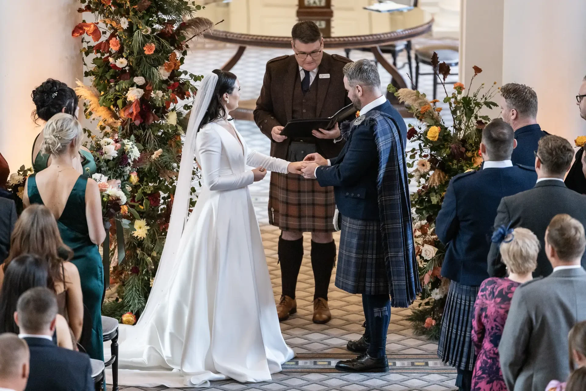 A bride and groom exchange vows, surrounded by guests and vibrant floral decorations in an elegant indoor setting, with the officiant and groom wearing traditional kilts.