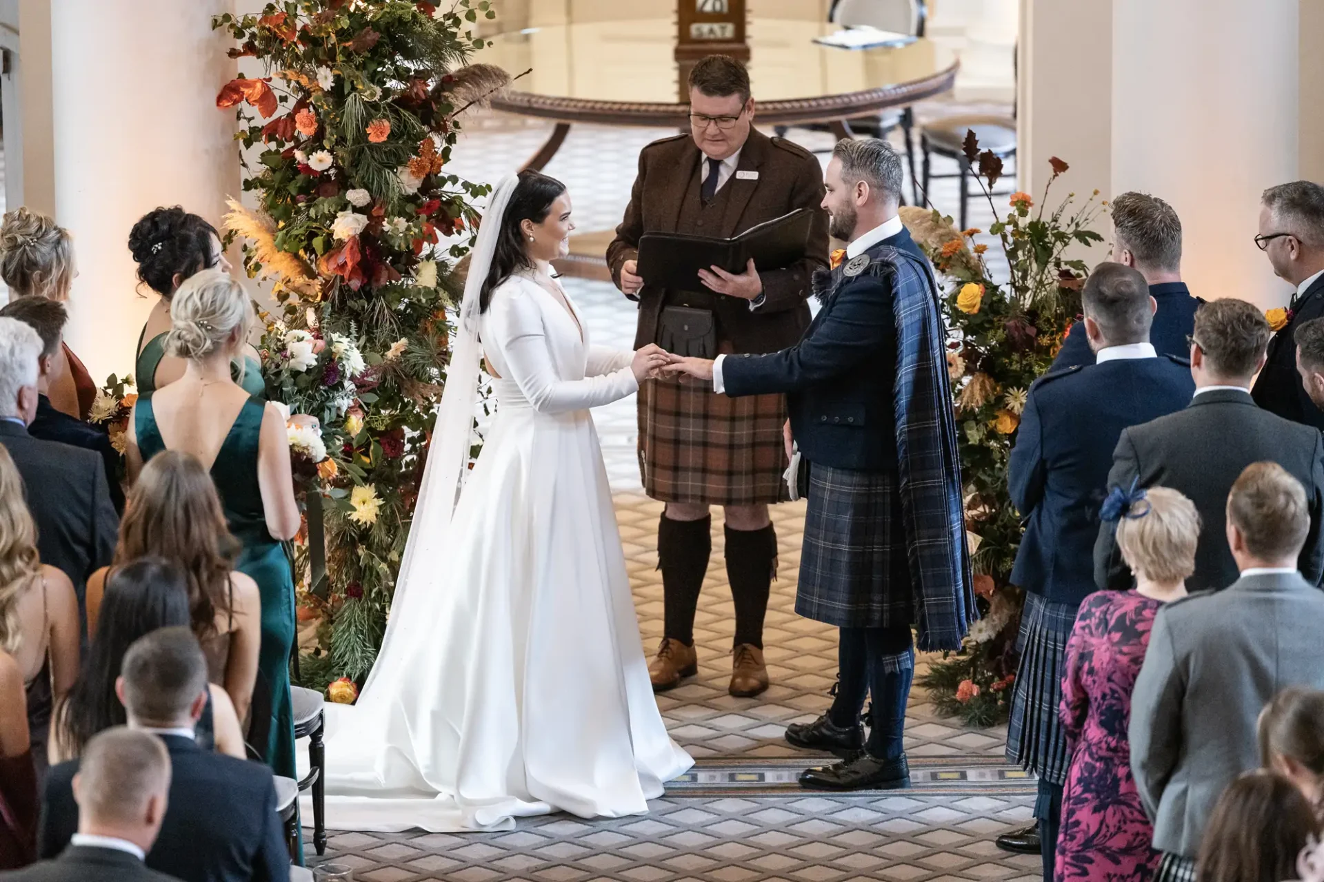 A bride and groom exchange vows in a wedding ceremony officiated by a man in a kilt, surrounded by guests and floral decorations in an elegant hall.