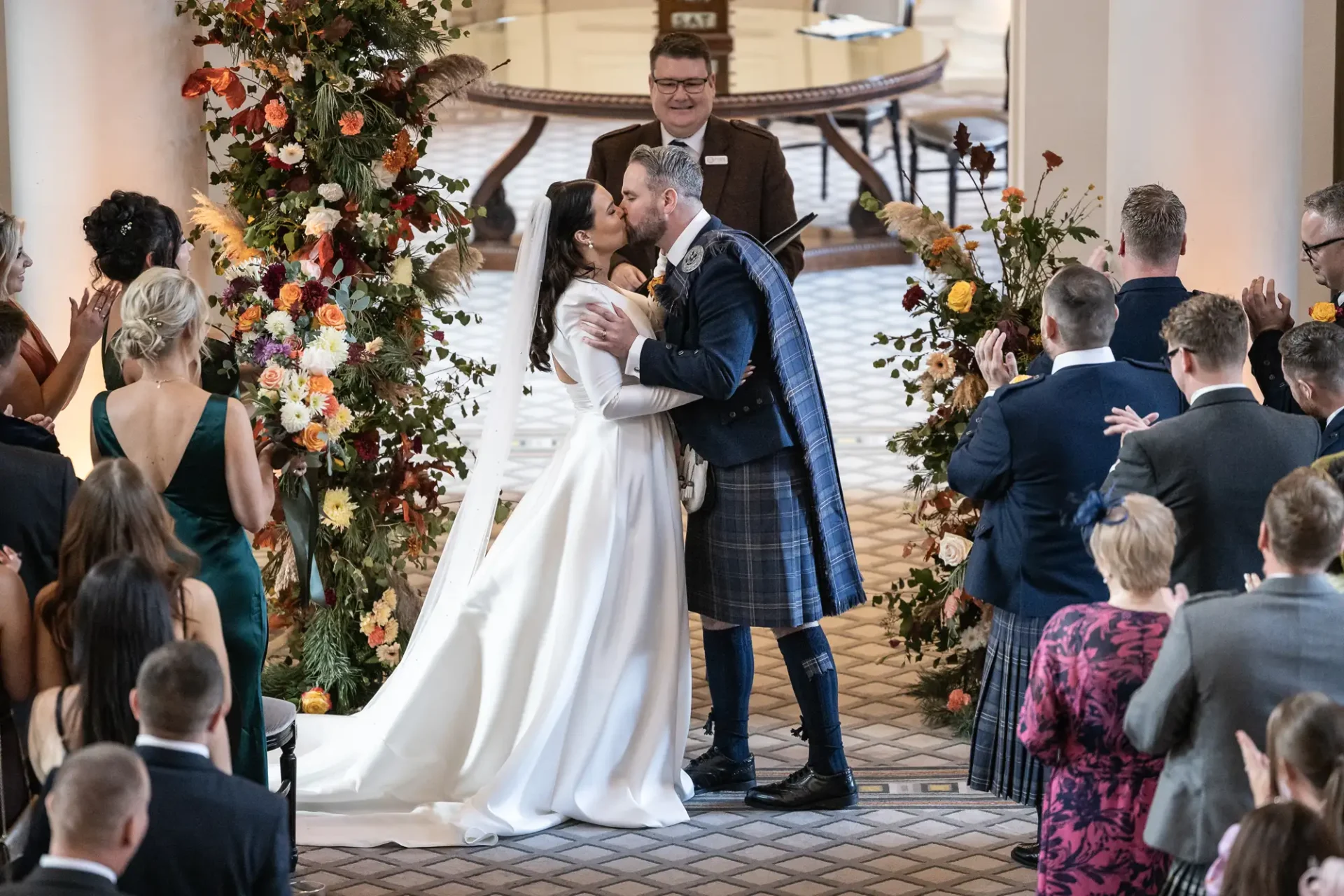 A bride and groom kiss at the altar, surrounded by guests and adorned with vibrant floral arrangements, with a celebrant watching in the background.
