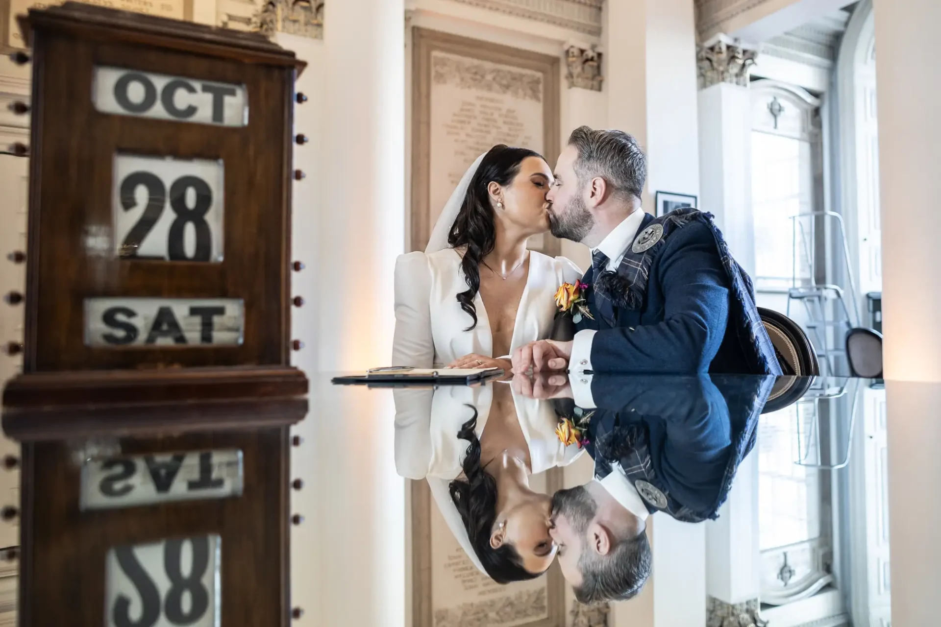 A bride and groom kissing, reflected in a glass table, with a nearby standing calendar showing the date october 28.