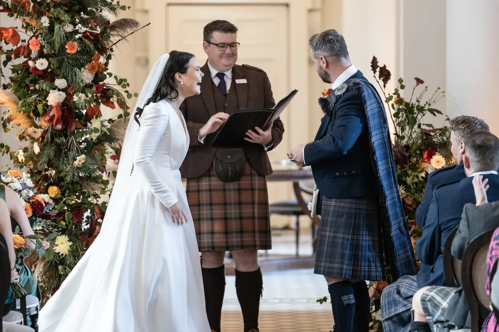 A bride in a white gown and a groom in a tartan kilt participate in a wedding ceremony indoors, officiated by a celebrant holding a book. audience members observe the event.