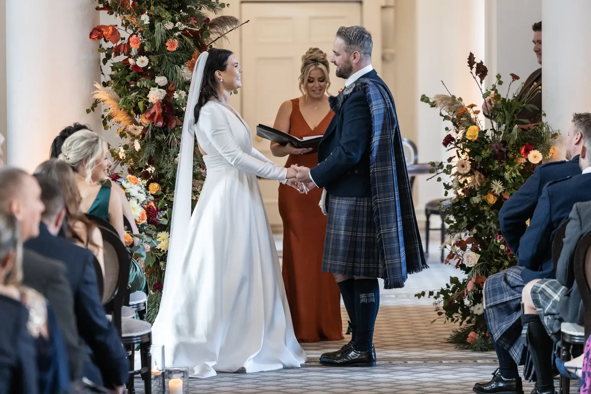 Bride and groom holding hands at their wedding ceremony, groom wearing a kilt, surrounded by guests and floral decorations.