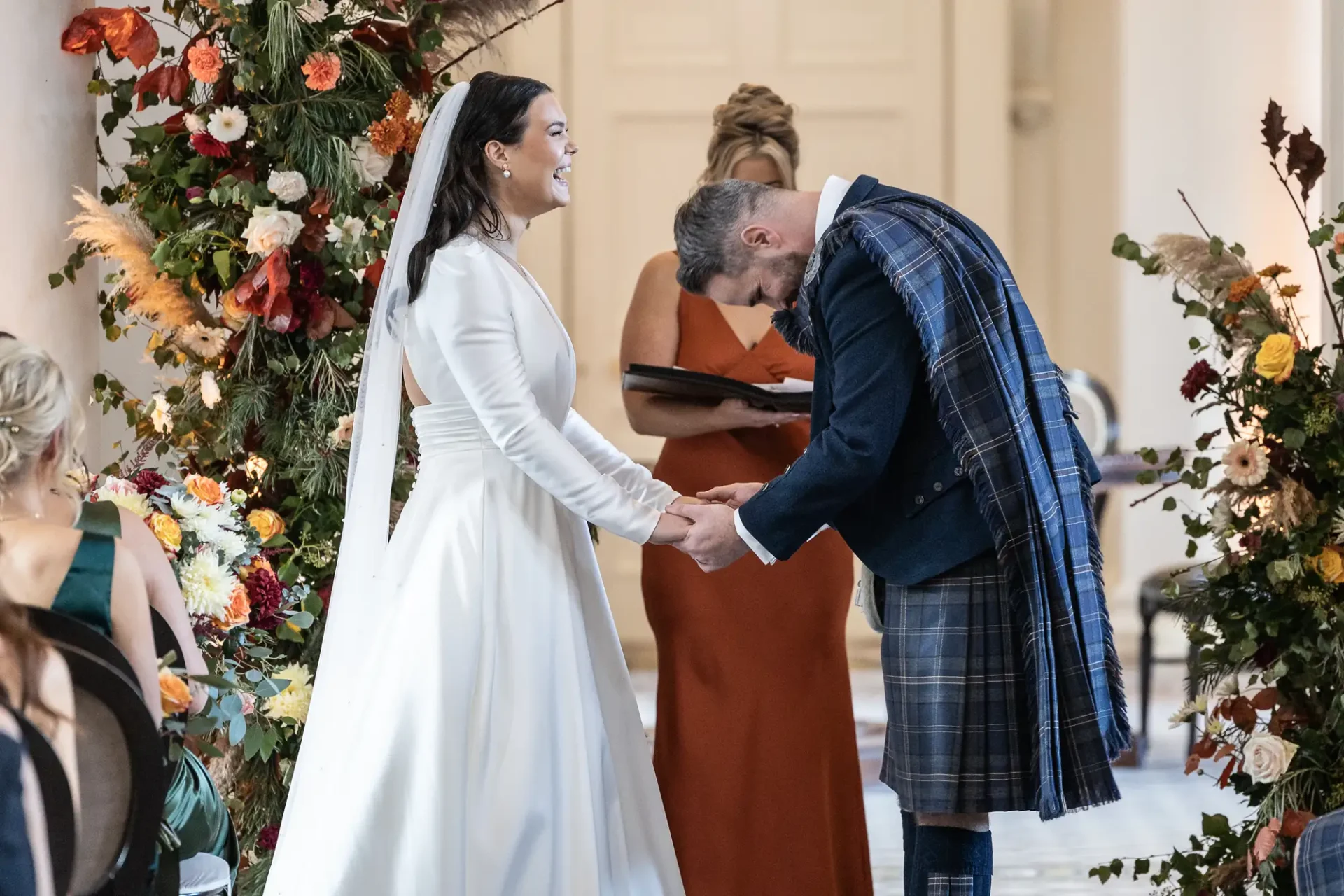 A groom in a kilt bows to a bride in a white dress at a floral-adorned wedding ceremony, with a bridesmaid in orange observing.