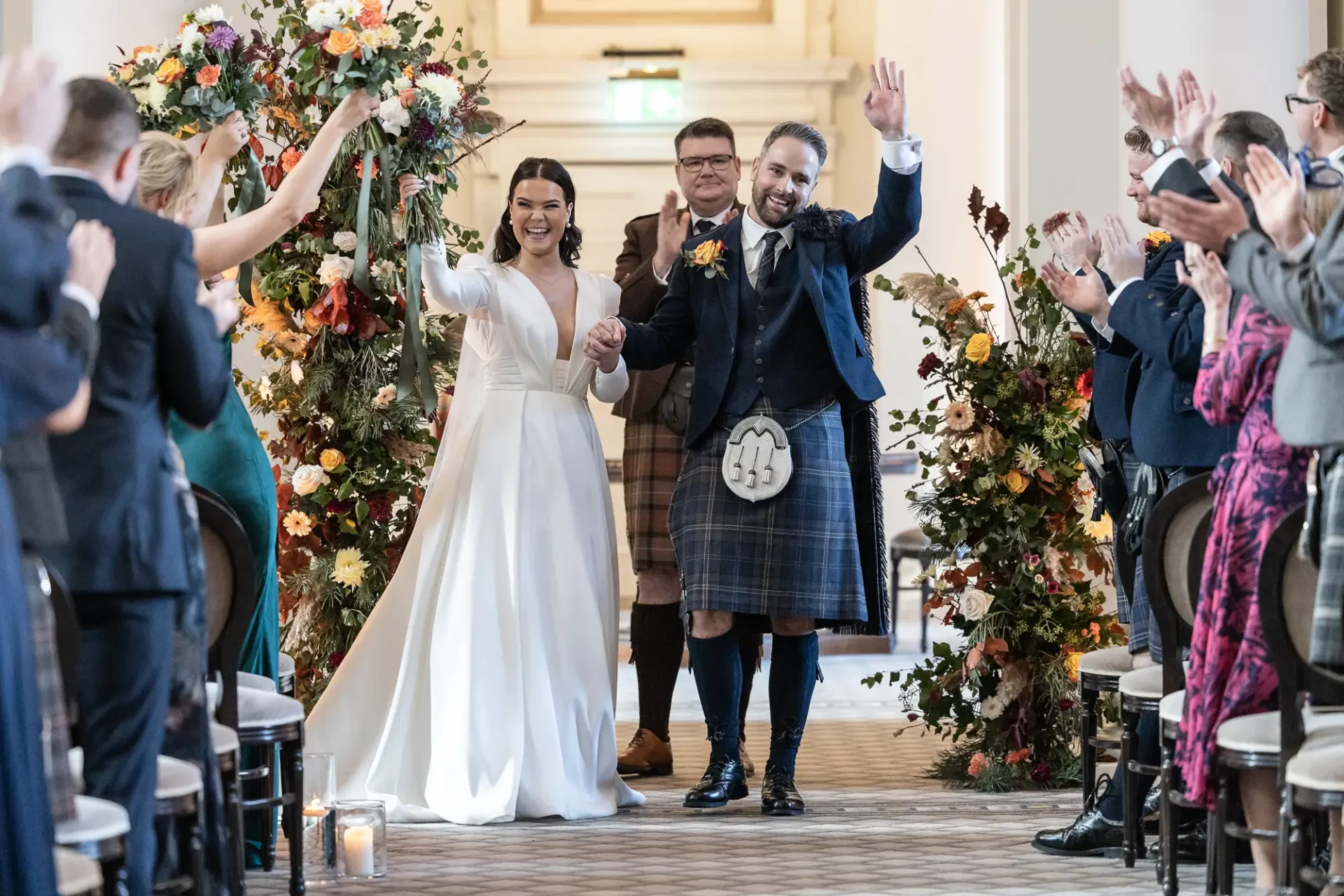 Bride in a white dress and groom in a kilt walk down the aisle, smiling, while guests applaud, surrounded by floral decorations.