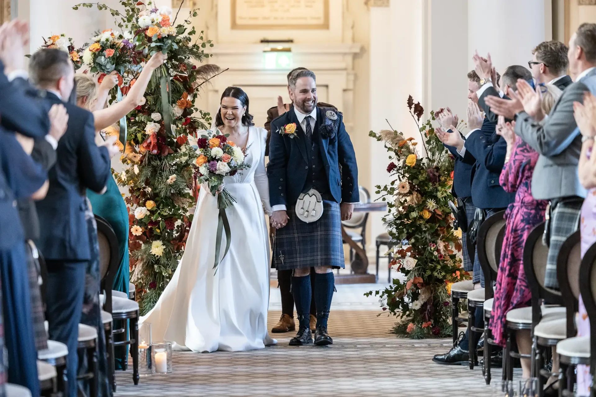 A joyful bride and groom walk down the aisle, receiving applause from guests, inside a decorated venue with floral archways. the groom wears a kilt.