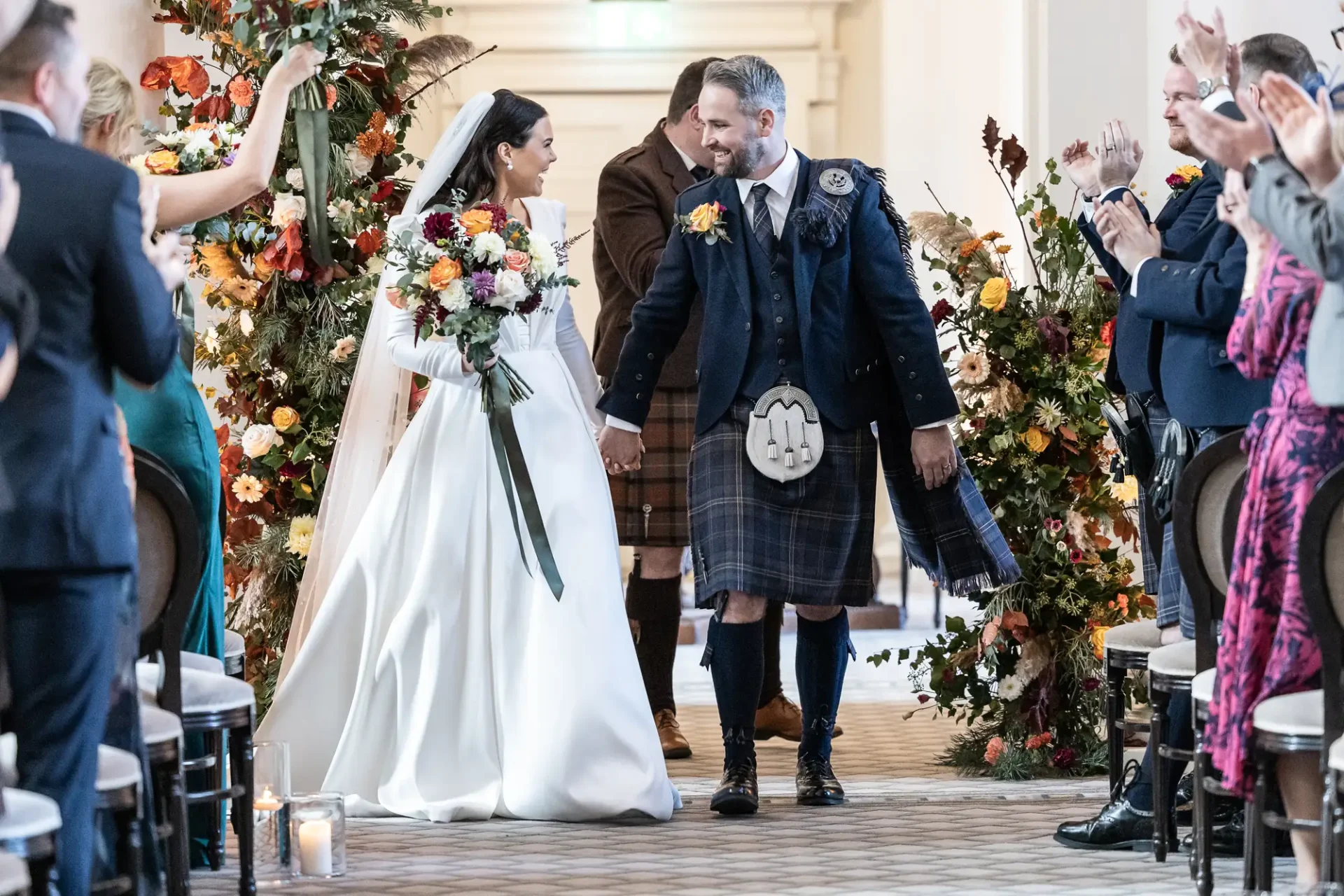 A newlywed couple walking down the aisle, receiving applause from guests, the bride in a white dress and the groom in a kilt.