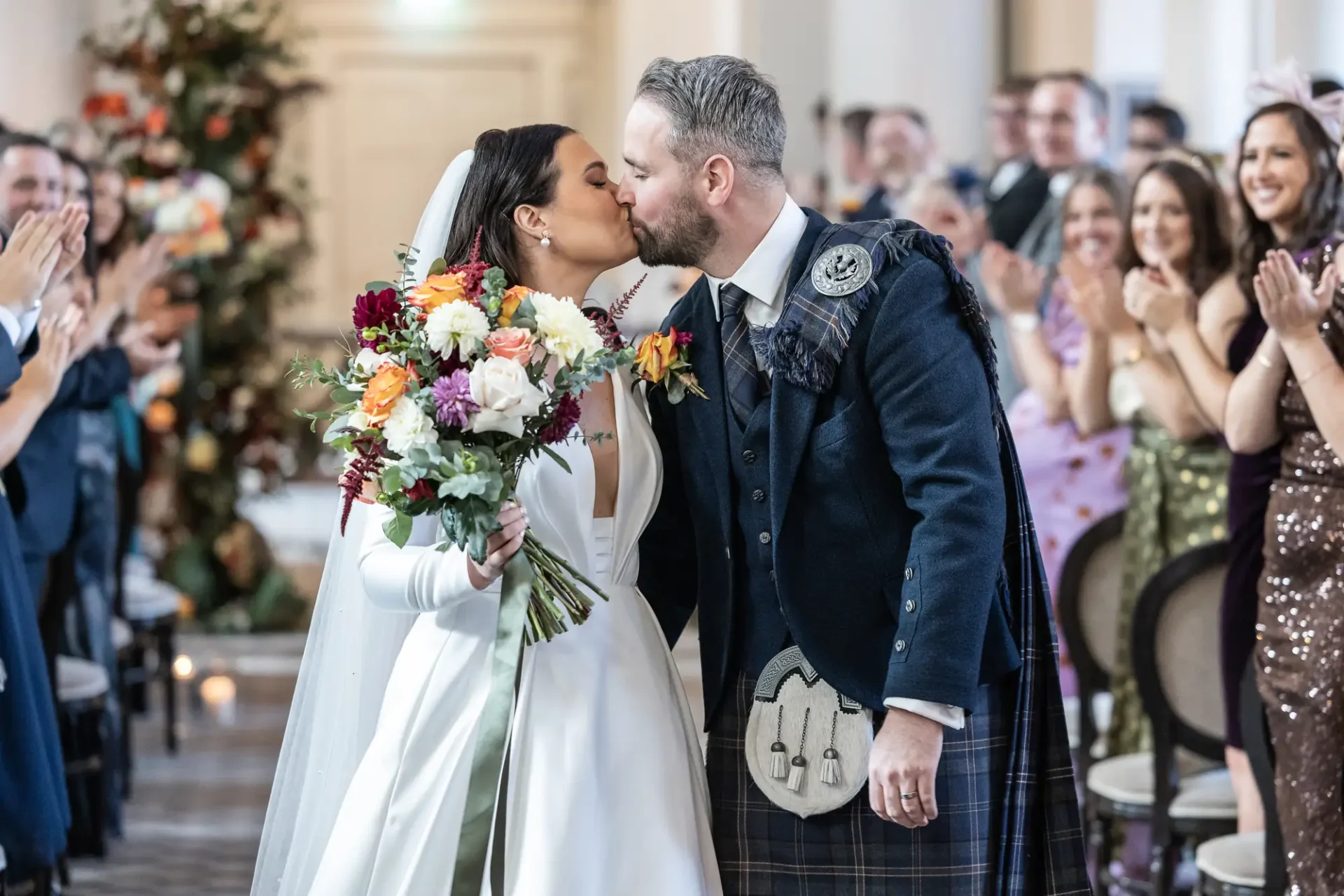 Bride and groom kissing, the groom in a kilt, at their wedding ceremony with guests clapping around them.