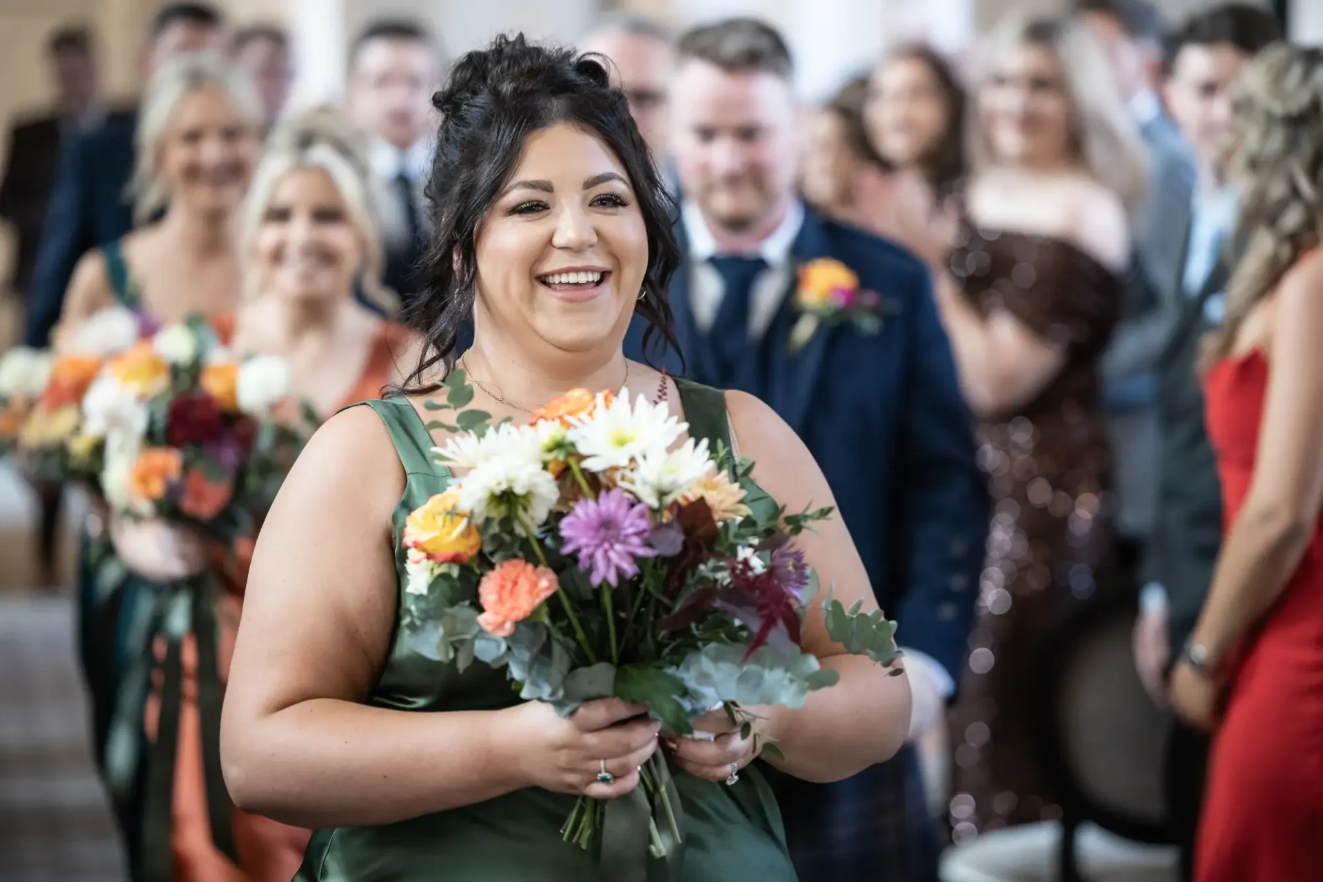 Bridesmaid in a green dress smiling as she walks down the aisle holding a bouquet, with wedding guests in the background.