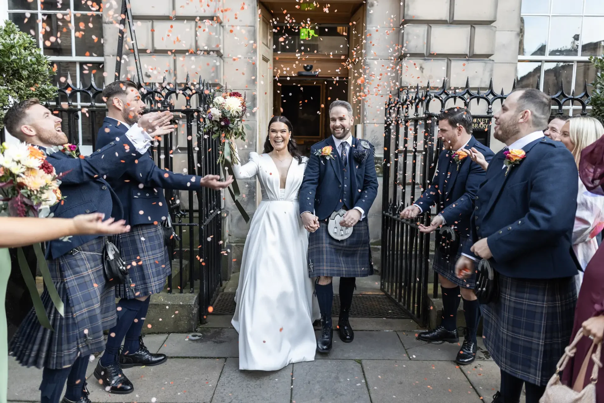 A newlywed couple exits a building, smiling as guests in kilts cheer and throw flower petals.