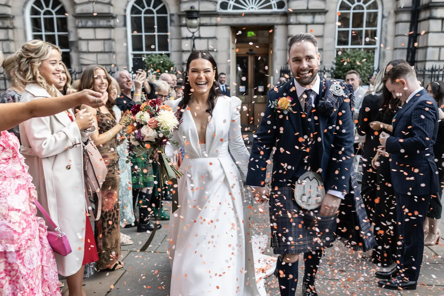 A bride in a white gown and a groom in a kilt smile while walking through a shower of confetti, surrounded by joyful guests outside a building.