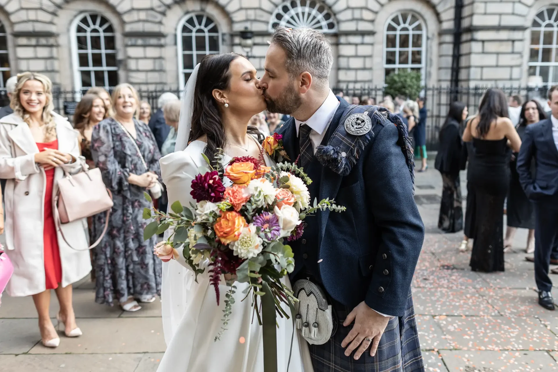 A newlywed couple kissing outside a building, surrounded by guests and confetti, with the groom wearing a kilt and the bride holding a bouquet.