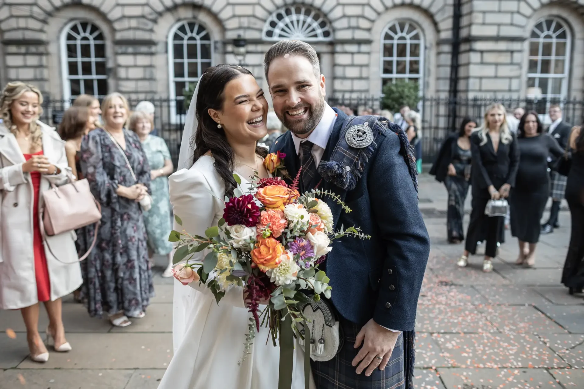A newlywed couple smiling outside a building, with the bride holding a bouquet and the groom wearing a kilt, surrounded by guests.