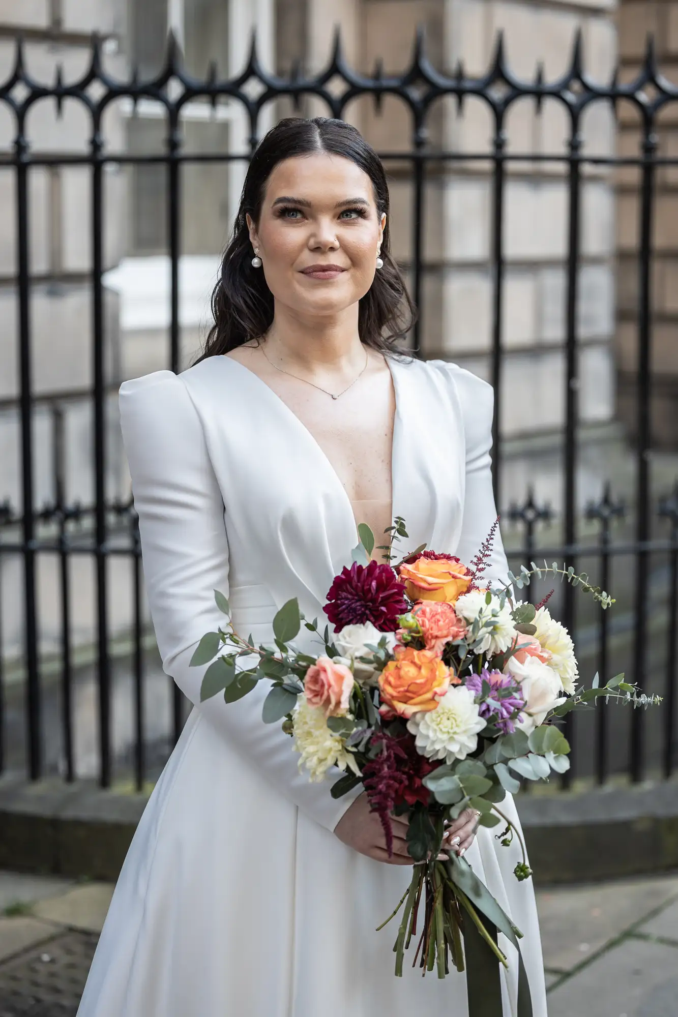 A bride in a white dress holds a colorful bouquet, smiling softly, with a historic building's fence in the background.